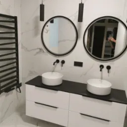 Nowoczesna łazienka z marmurowymi płytkami, podwójną umywalką nablatową na białej szafce z czarnym blatem, czarnymi okrągłymi lustrami i grzejnikiem drabinkowym w kolorze czarnym, oraz wiszącymi...