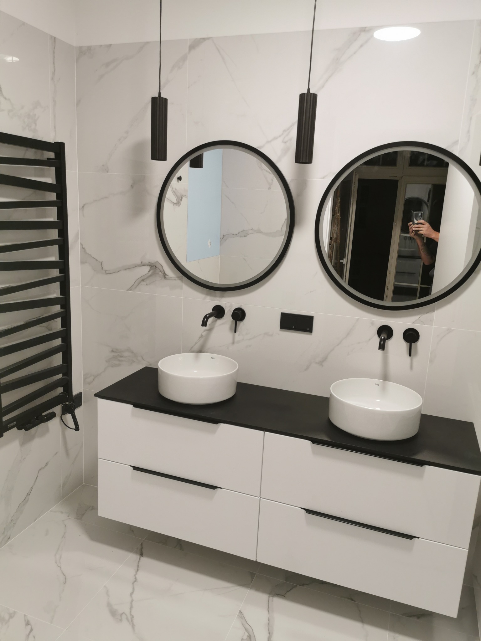 Nowoczesna łazienka z marmurowymi płytkami, podwójną umywalką nablatową na białej szafce z czarnym blatem, czarnymi okrągłymi lustrami i grzejnikiem drabinkowym w kolorze czarnym, oraz wiszącymi...