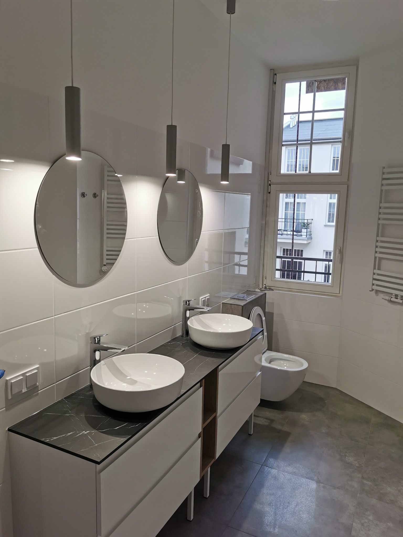 Nowoczesna łazienka z dwoma okrągłymi lustrami, białymi umywalkami na szafce z czarnym blatem imitującym marmur, podwieszaną toaletą i widokiem na balkon zza okna.