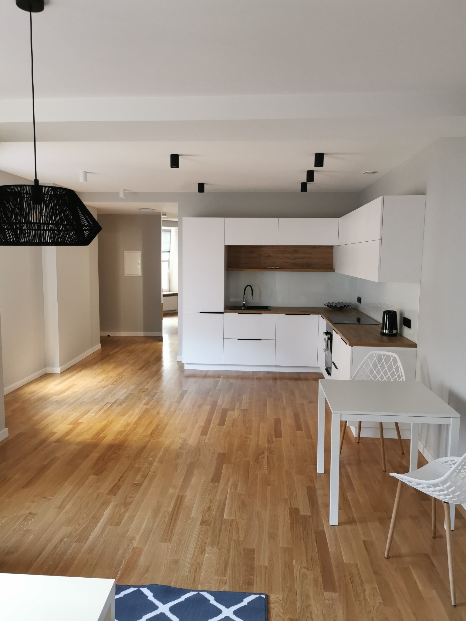 Jasne, przestronne wnętrze z drewnianą podłogą, białą kuchnią z drewnianymi akcentami, stołem i krzesłami, oświetlone wiszącą lampą z czarnego rattanu i punktowymi reflektorami.