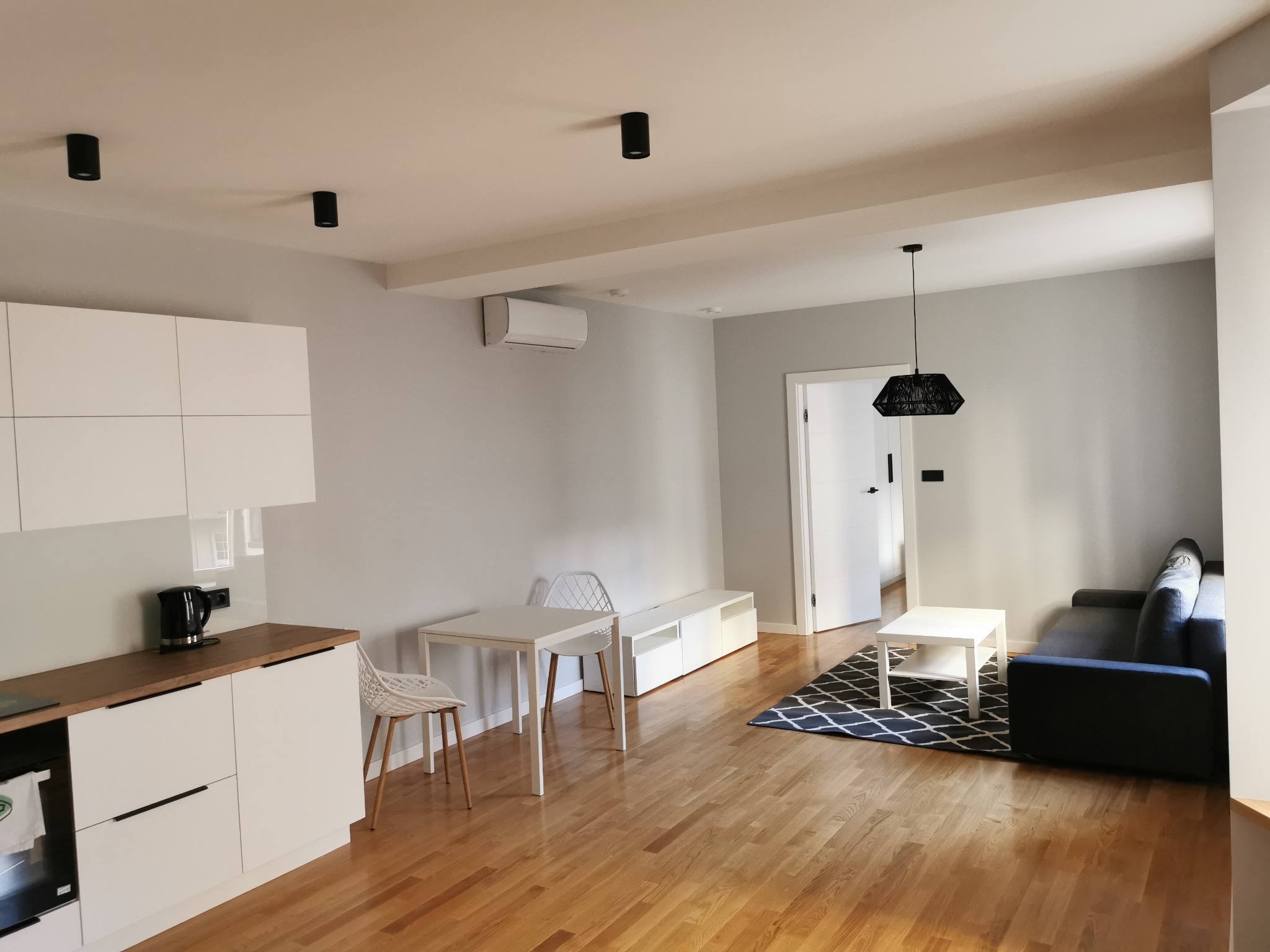 Jasne, przestronne wnętrze mieszkania z parkietem, obejmujące aneks kuchenny z białymi szafkami i drewnianym blatem, stół z krzesłami, sofę i geometryczny dywan, oświetlone czarnymi lampami...