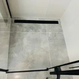 Szare płytki podłogowe z odpływem liniowym w kolorze czarnym, kabina prysznicowa z czarnymi profilami, białe płytki ścienne.