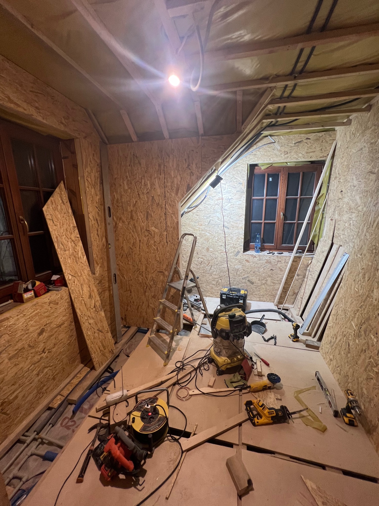 Wnętrze w trakcie remontu: ściany i sufit obłożone płytami OSB, widoczne okna z drewnianą stolarką, drabina, narzędzia (wiertarka, piła szablasta, odkurzacz przemysłowy) oraz instalacje elektryczne...