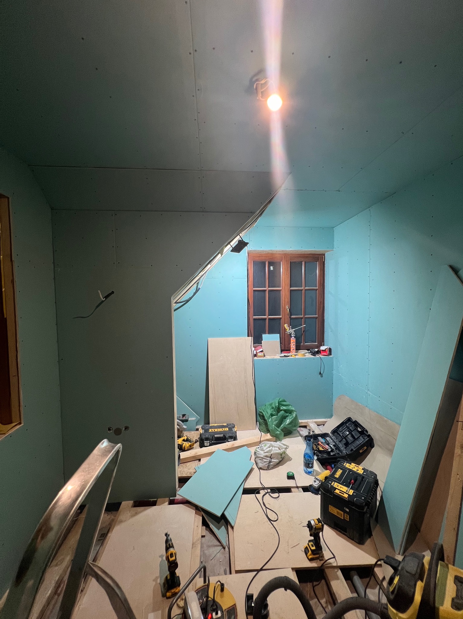 Pokój na poddaszu w trakcie montażu płyt gipsowo-kartonowych, widoczne narzędzia, skrzynki narzędziowe i okno z drewnianą ramą.