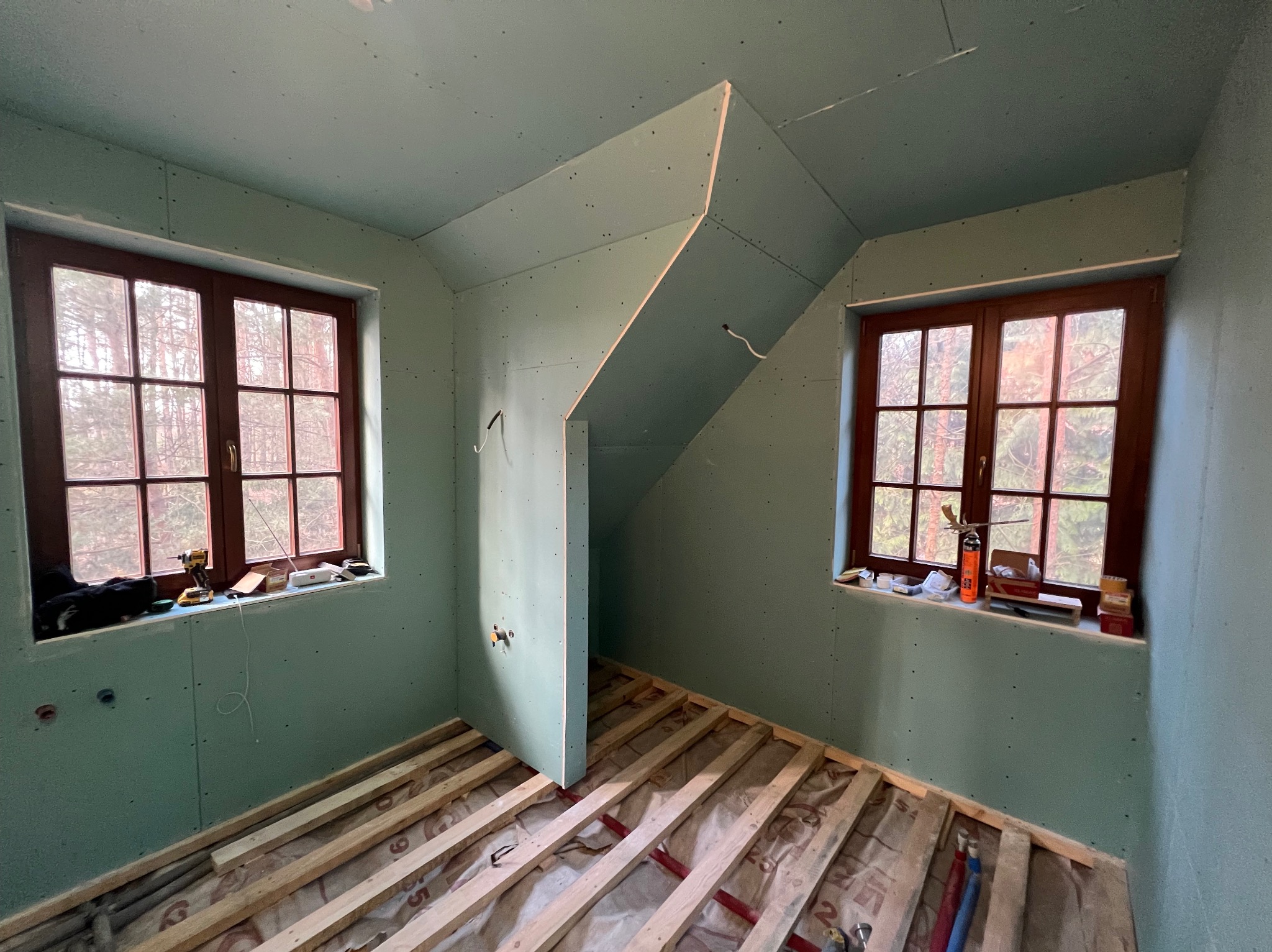 Pokój na poddaszu w trakcie prac wykończeniowych, z płytami gipsowo-kartonowymi na ścianach i suficie oraz drewnianą konstrukcją podłogi, widok na drzewa za oknami.