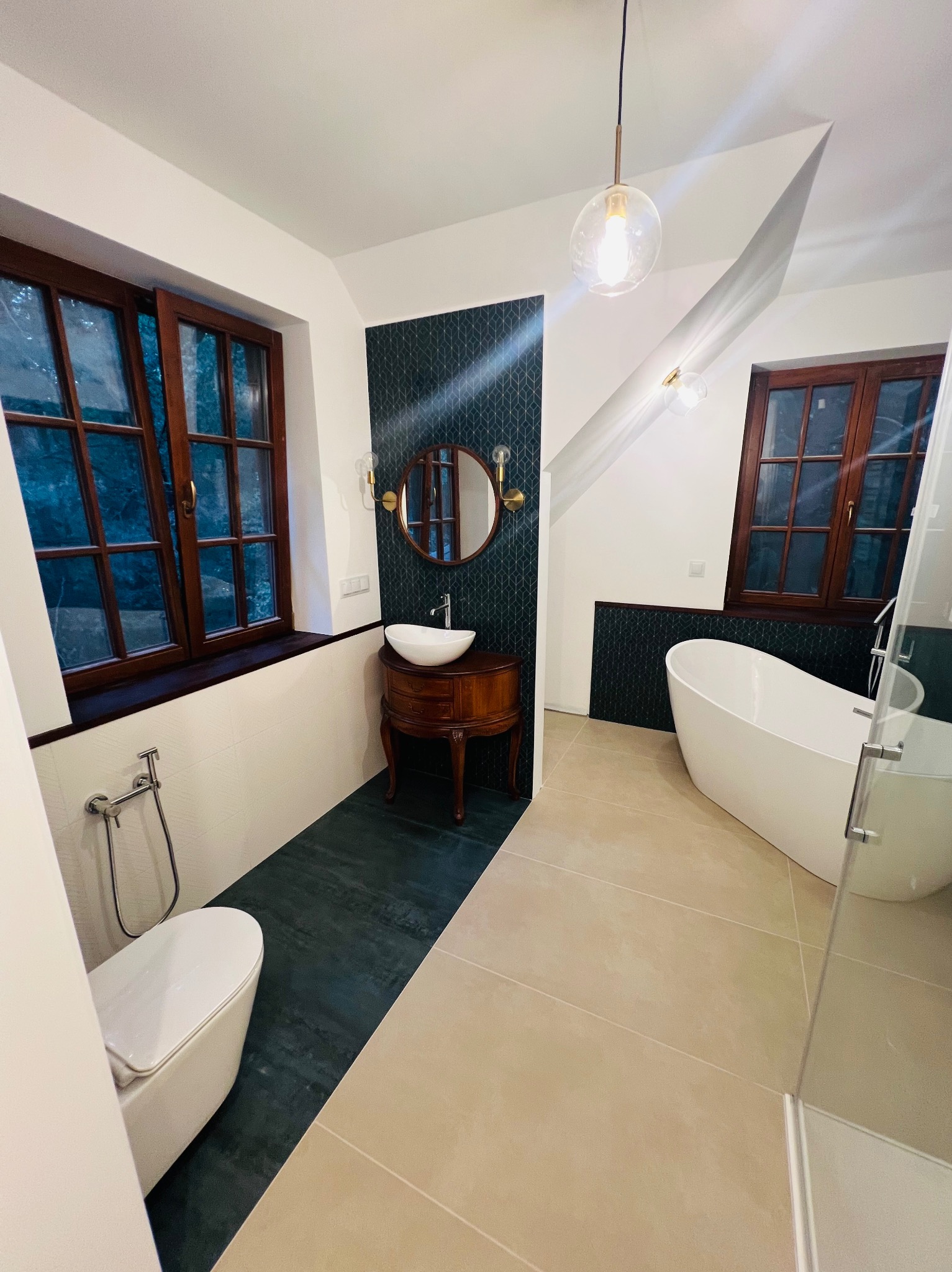 Nowoczesna łazienka z wolnostojącą wanną, umywalką na drewnianej szafce w stylu retro, oknami w drewnianych ramach i podłogą z płytek w dwóch odcieniach.