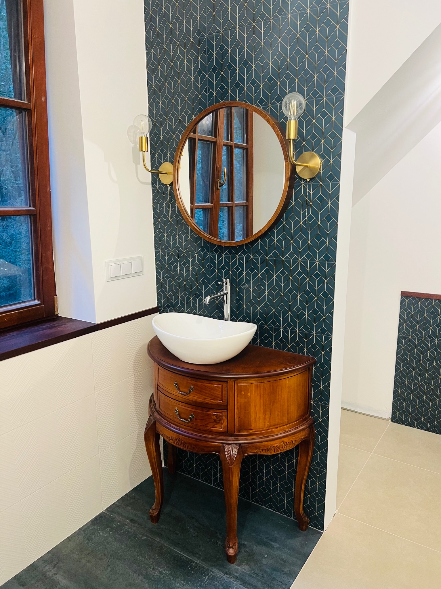 Okrągłe lustro w drewnianej ramie nad umywalką nablatową na szafce w stylu retro, geometryczna tapeta w odcieniach granatu i złota, złote kinkiety po bokach lustra.