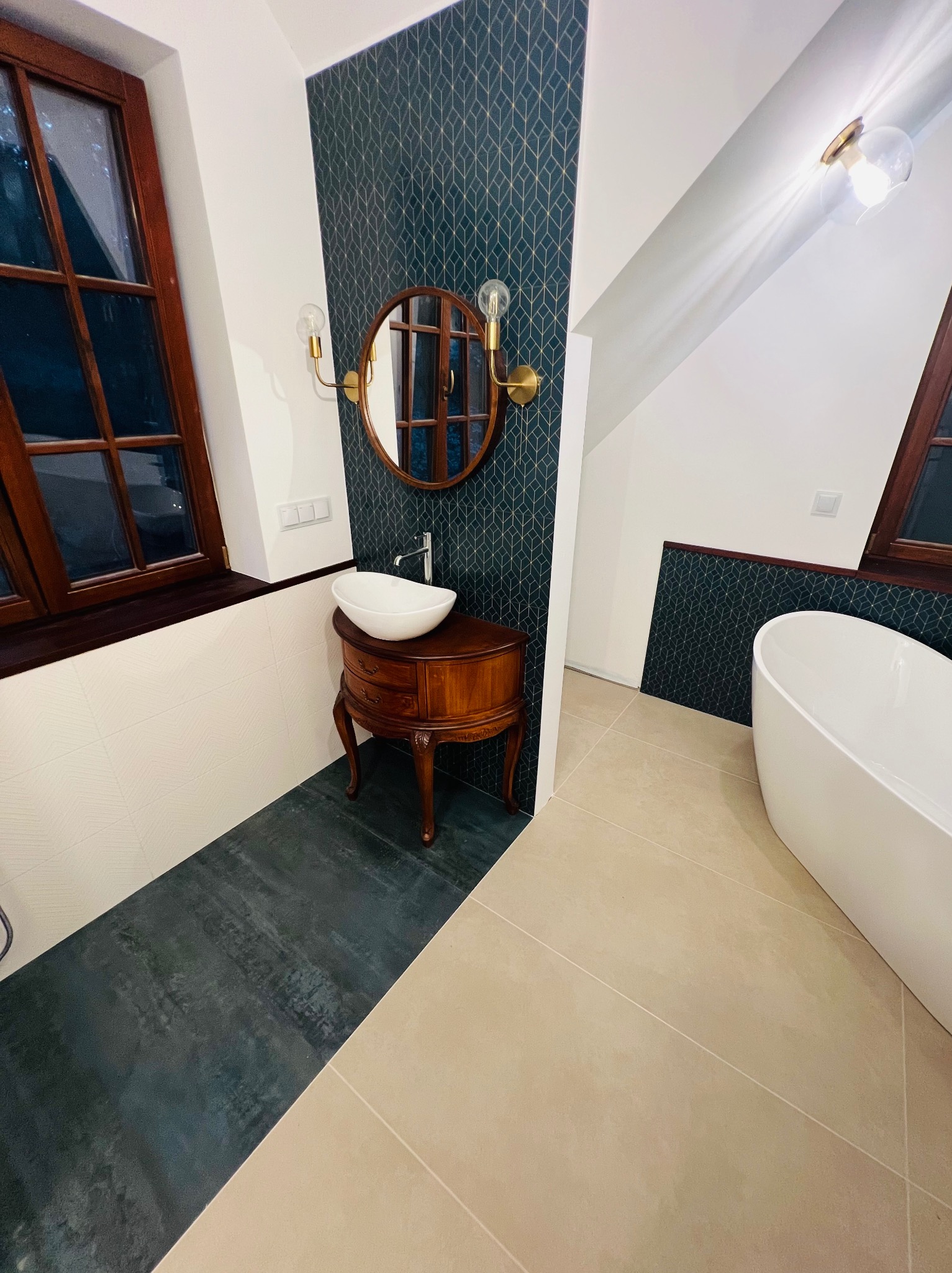 Wykończona łazienka w stylu retro: drewniana szafka z umywalką nablatową, okrągłe lustro z oświetleniem, ciemna ściana z geometrycznym wzorem, biała wanna wolnostojąca i jasna podłoga z fugami.