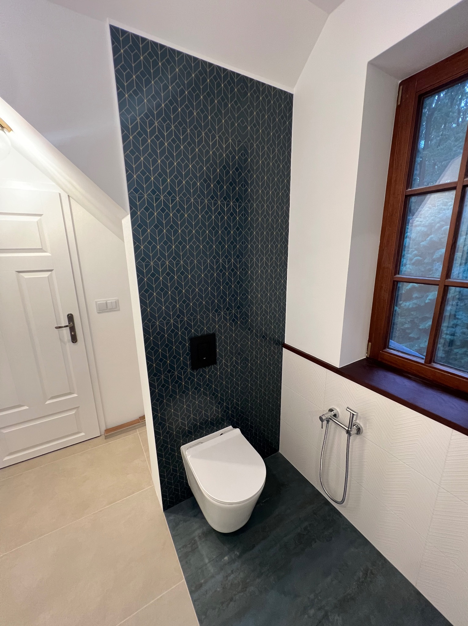 Nowoczesna łazienka z toaletą podwieszaną, ciemną geometryczną tapetą na jednej ścianie, białą płytką z fakturą na innej, drewnianą ramą okienną i bidetką