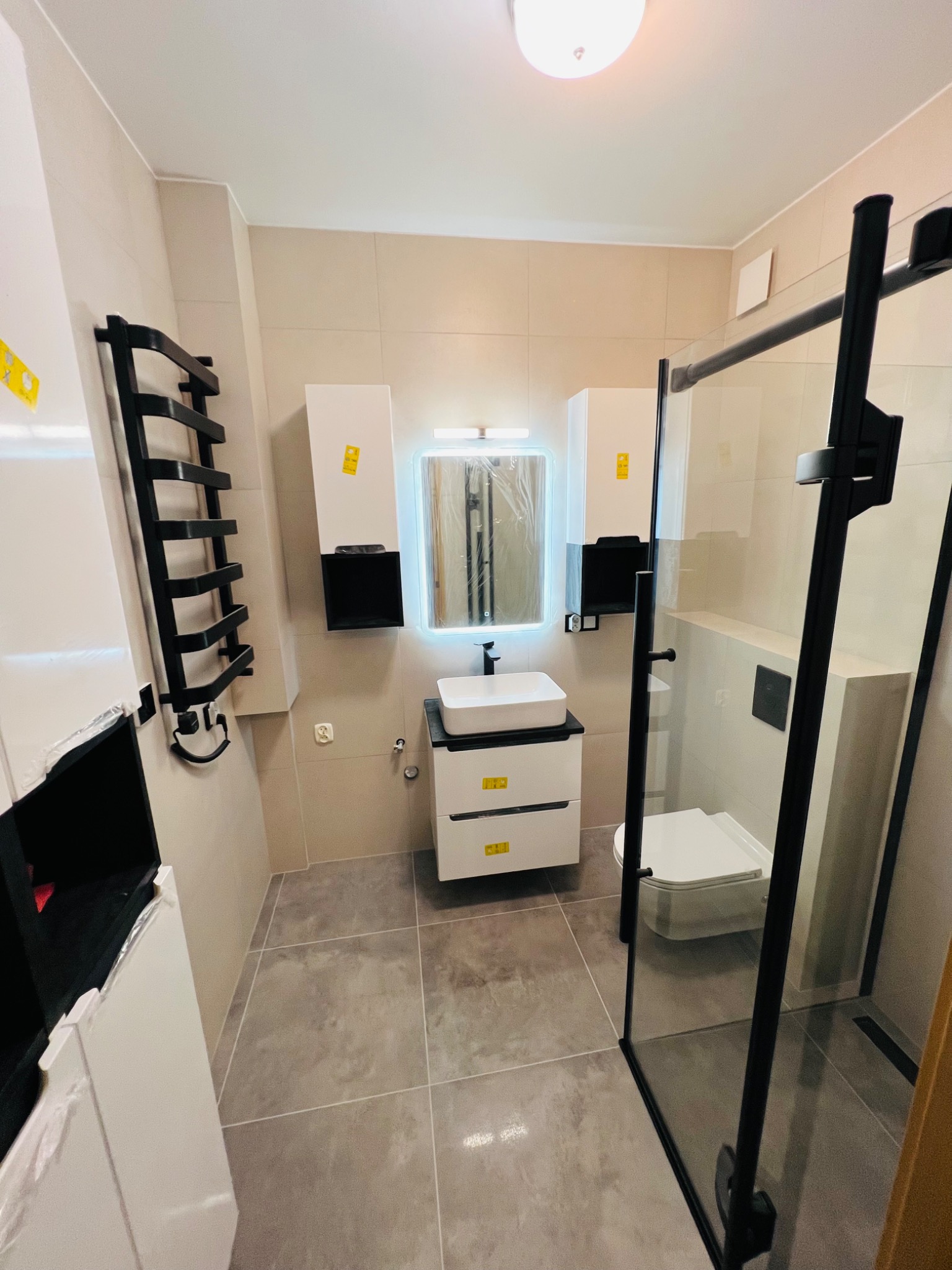 Nowoczesna łazienka z kabiną prysznicową typu walk-in z czarnymi ramami, umywalką nablatową na szafce z szufladami, podświetlanym lustrem i grzejnikiem drabinkowym.