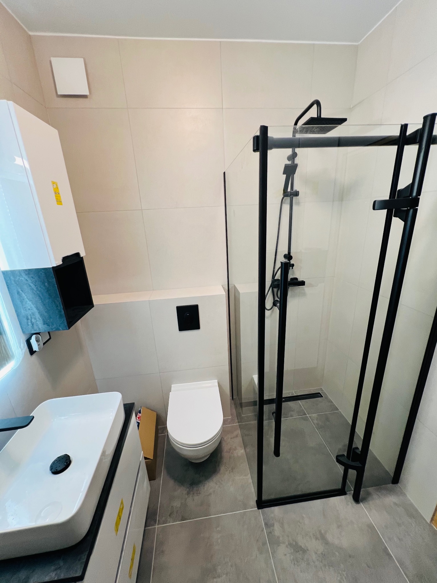 Nowoczesna łazienka z kabiną prysznicową typu walk-in z czarnymi profilami, podwieszaną toaletą i umywalką nablatową na szafce z szufladami, szare płytki podłogowe i ścienne.