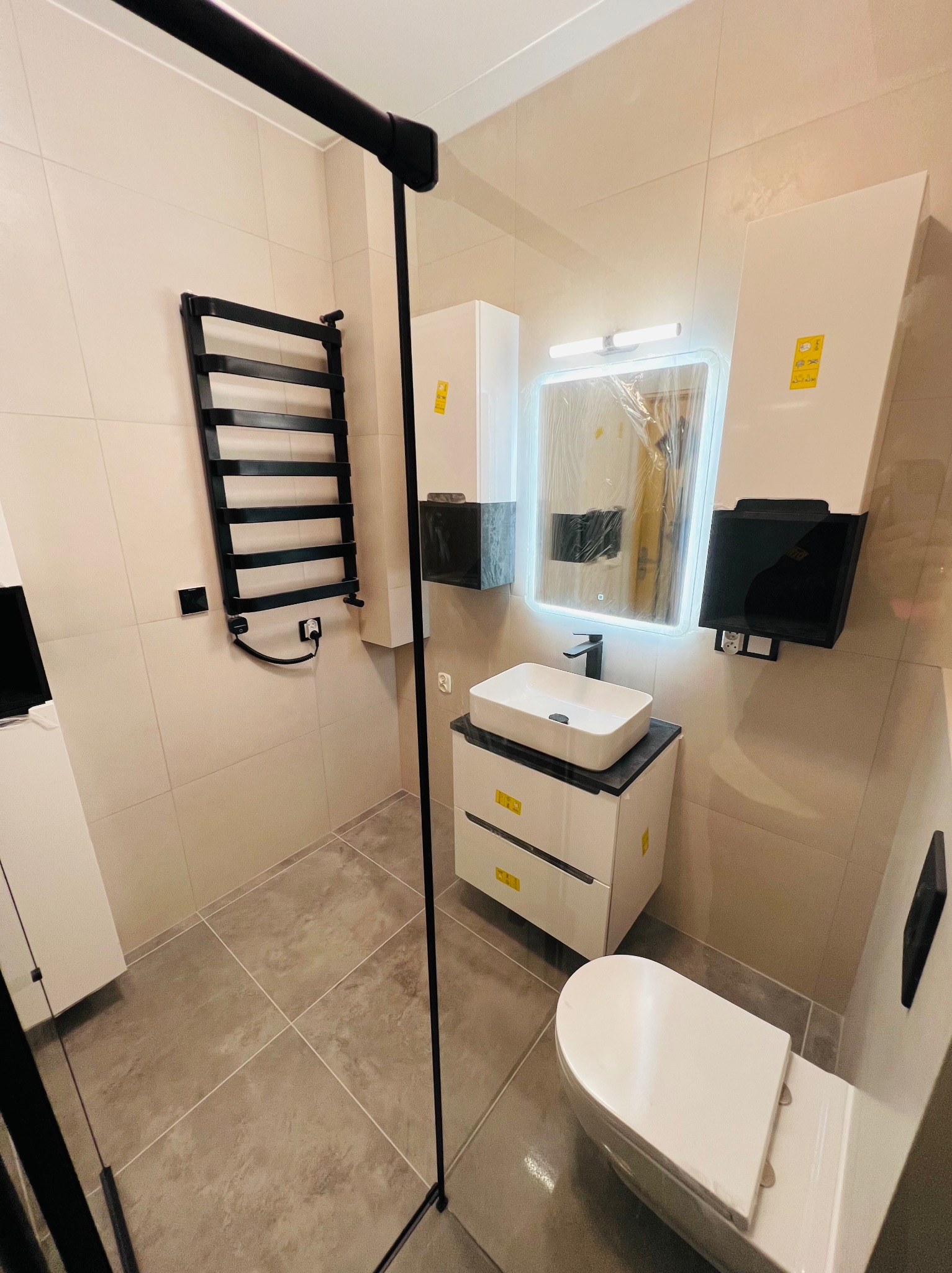 Nowoczesna łazienka z beżowymi płytkami, czarnym grzejnikiem drabinkowym, umywalką nablatową na szafce z szufladami oraz podświetlanym lustrem, częściowo jeszcze w folii ochronnej.