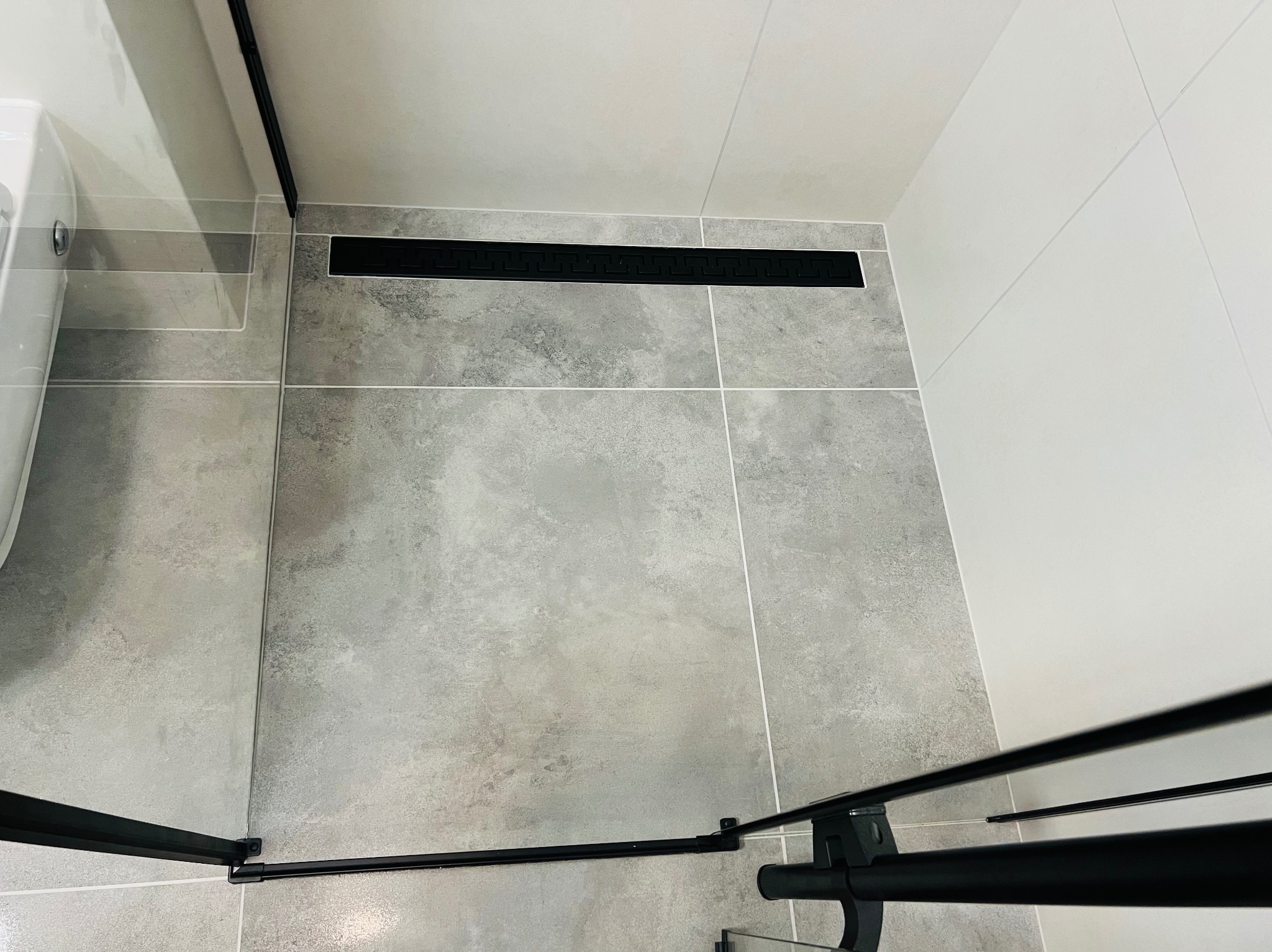 Szare płytki podłogowe z odpływem liniowym w kolorze czarnym, kabina prysznicowa z czarnymi profilami, białe płytki ścienne.