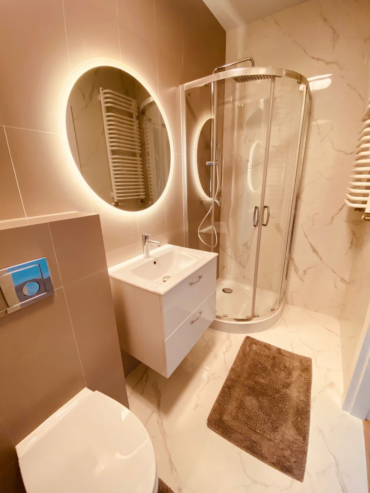 Nowoczesna łazienka z owalnym lustrem podświetlanym LED, kabiną prysznicową z zaokrąglonymi szybami i marmurową posadzką, wykończona w beżowej kolorystyce.