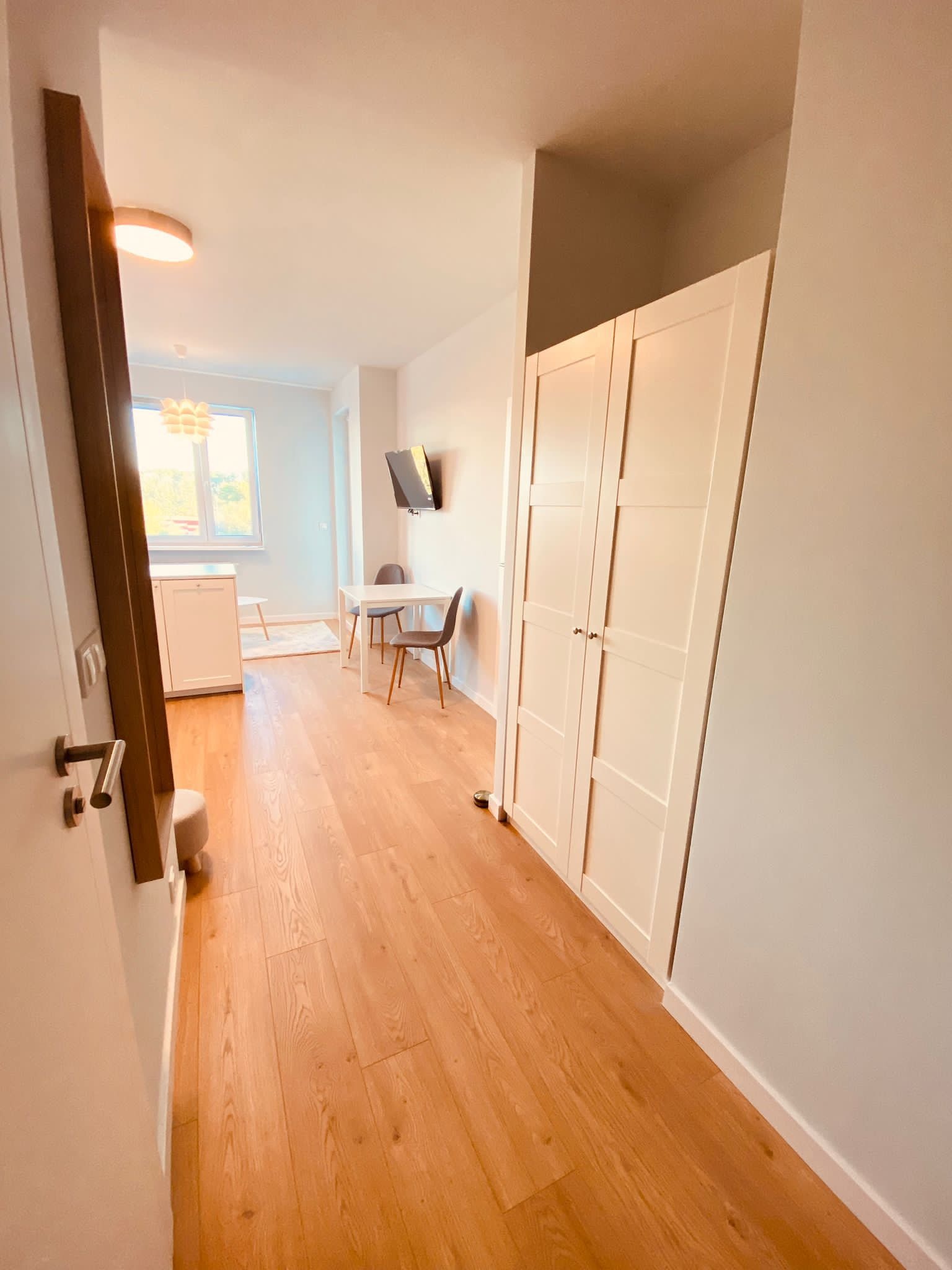 Jasne wnętrze mieszkania z drewnianą podłogą, białymi ścianami i szafą wnękową oraz widokiem na aneks kuchenny z jadalnią, oświetlone naturalnym światłem.