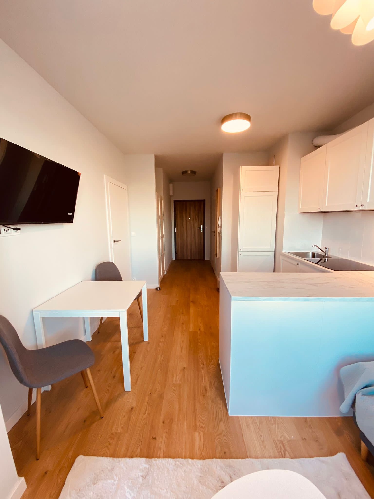 Jasne wnętrze mieszkania z drewnianą podłogą, białymi meblami kuchennymi i stołem z krzesłami, widok na przedpokój z drewnianymi drzwiami.