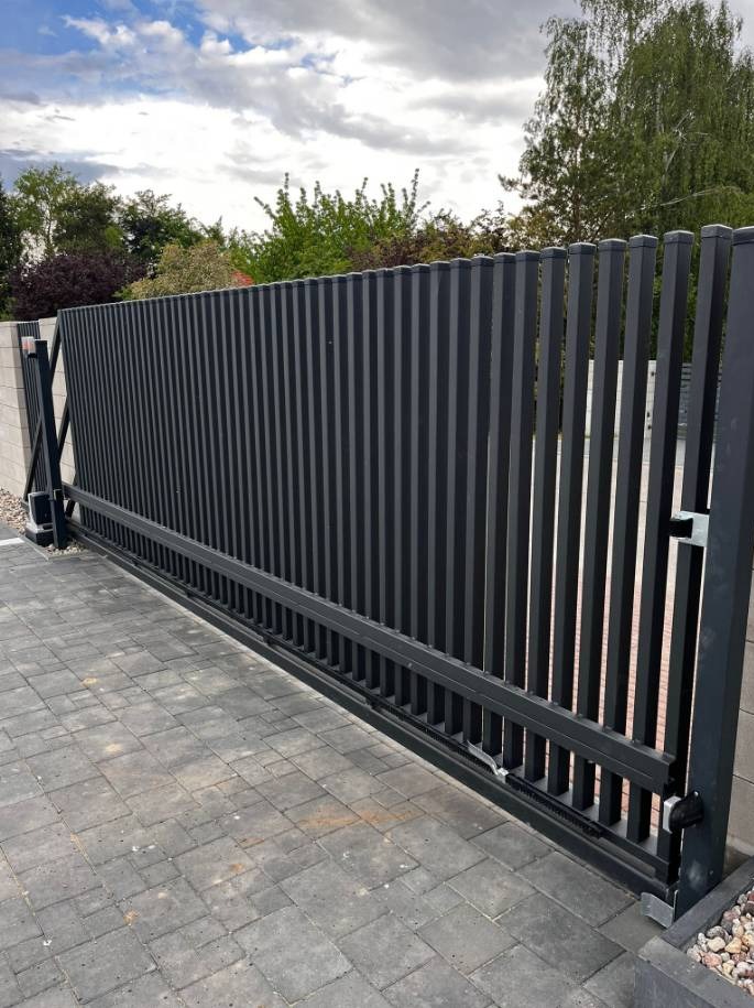 Nowoczesna, przesuwna brama w kolorze antracytu z pionowych profili, widoczny mechanizm jezdny i kostka brukowa na podjeździe.