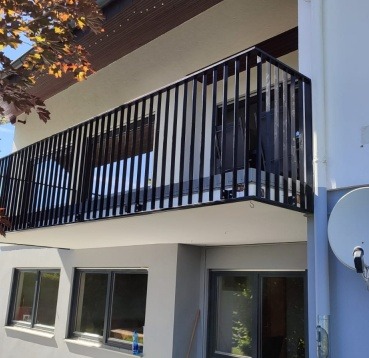 Czarna, metalowa balustrada balkonowa z pionowymi szczeblami, zamontowana na jasnym balkonie budynku mieszkalnego z widocznymi oknami i fragmentem drzewa w tle.