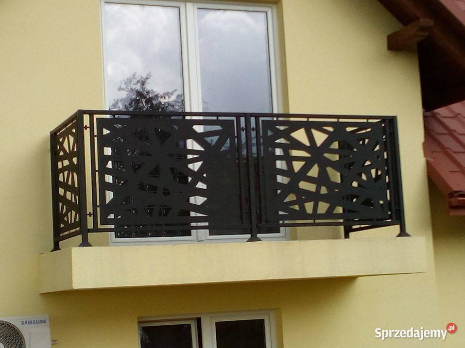 Czarna, metalowa balustrada balkonowa z geometrycznym, ażurowym wzorem na tle żółtej elewacji budynku z białymi oknami.