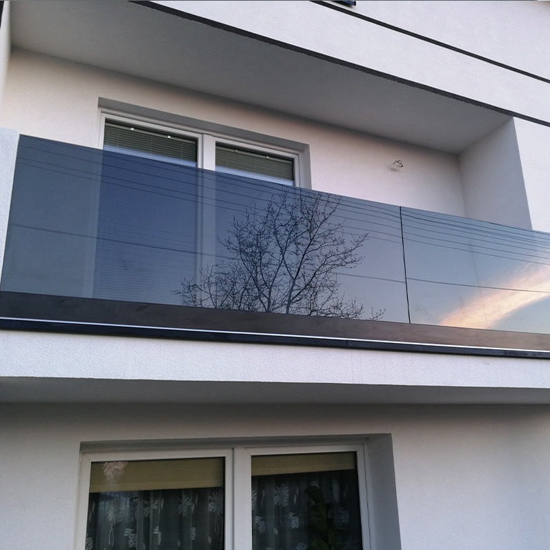 Szklana balustrada balkonowa z widocznym odbiciem drzewa i zachodzącego słońca, zamontowana na białym budynku z oknami.