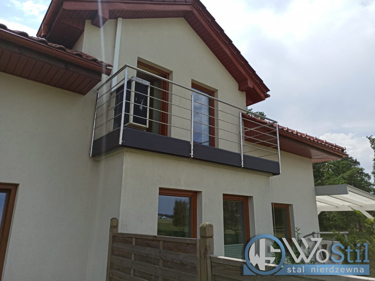 Balkon z nowoczesną, poziomą balustradą ze stali nierdzewnej, zamontowaną na budynku z beżową elewacją i brązowymi ramami okiennymi. Na balkonie stoi klimatyzator. W tle widoczny fragment ogrodzenia.