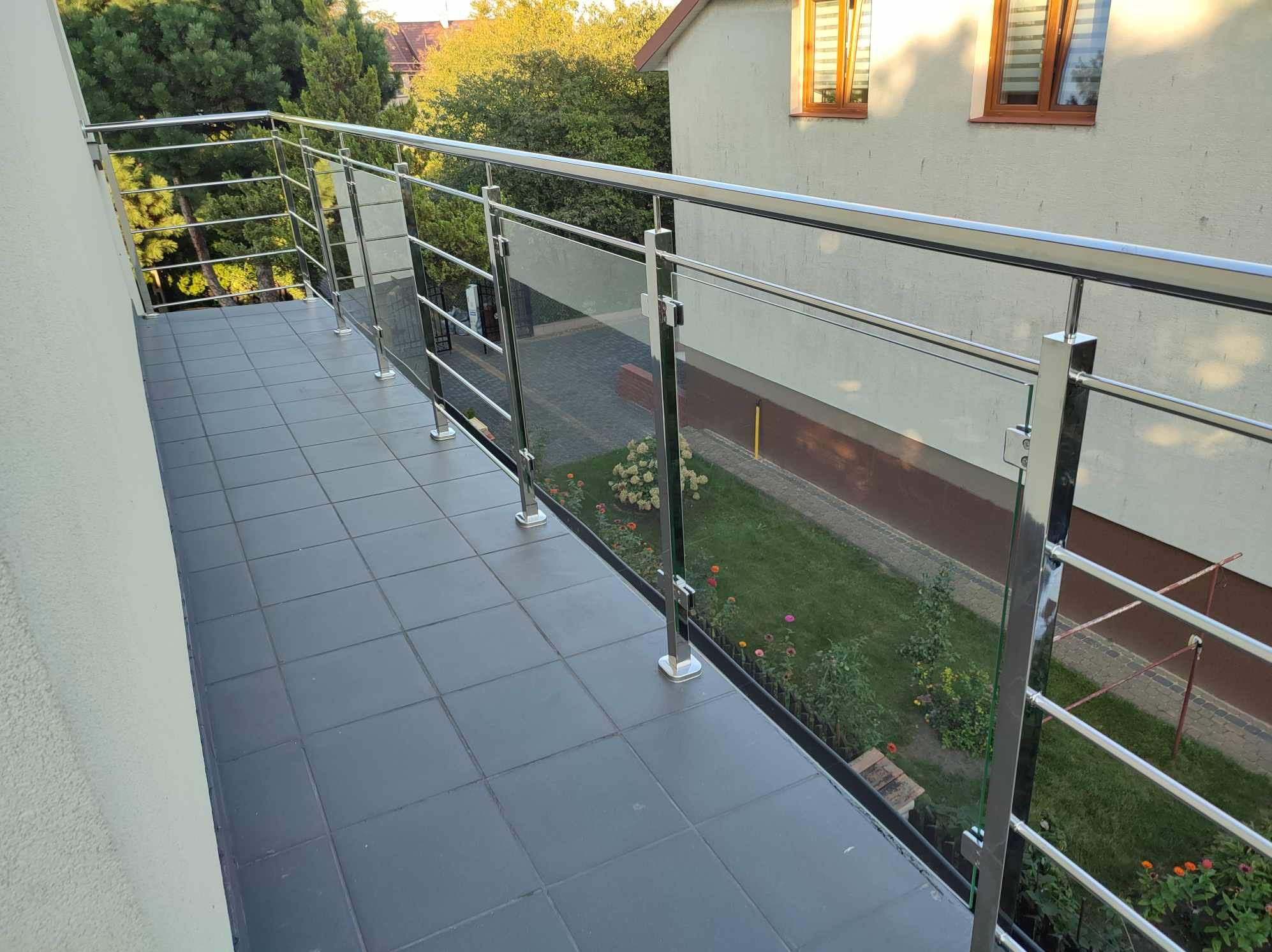 Balkon z nowoczesną balustradą ze stali nierdzewnej i szkła, widok na ogród z kwitnącymi kwiatami i trawnikiem, szare płytki na podłodze.