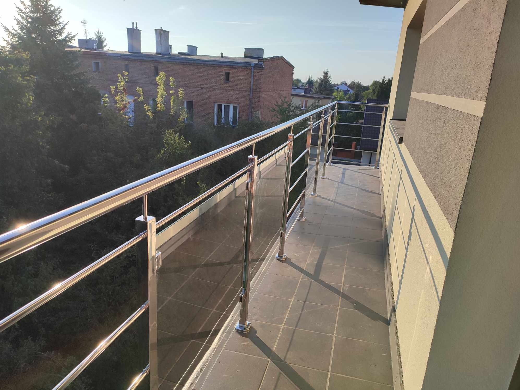 Balkon z nowoczesną balustradą ze stali nierdzewnej i szkła, widok na budynek mieszkalny i zieleń w słoneczny dzień.