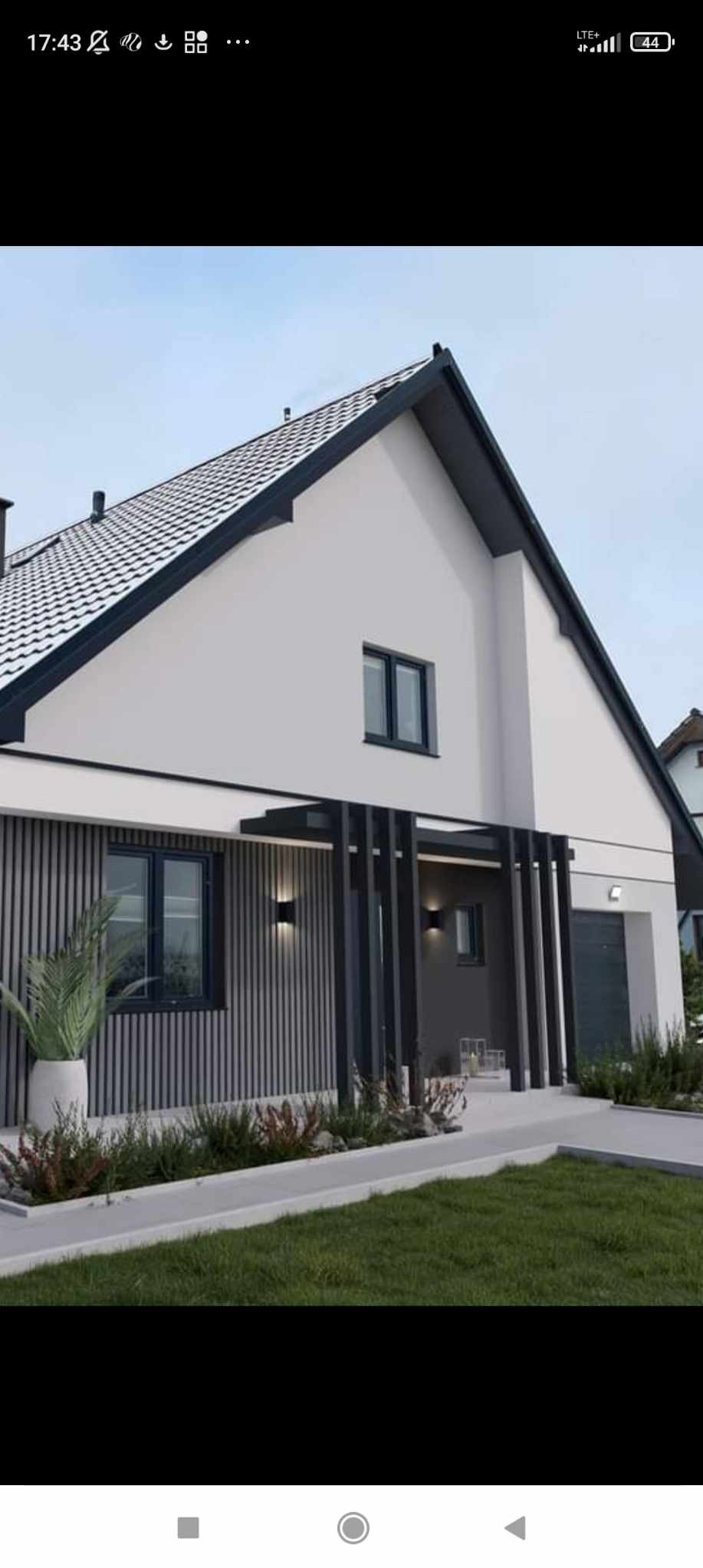 Nowoczesny dom jednorodzinny z czarnymi oknami PCV, elewacją w odcieniach bieli i szarości oraz drewnianą okładziną pionową, oświetlony lampami zewnętrznymi.