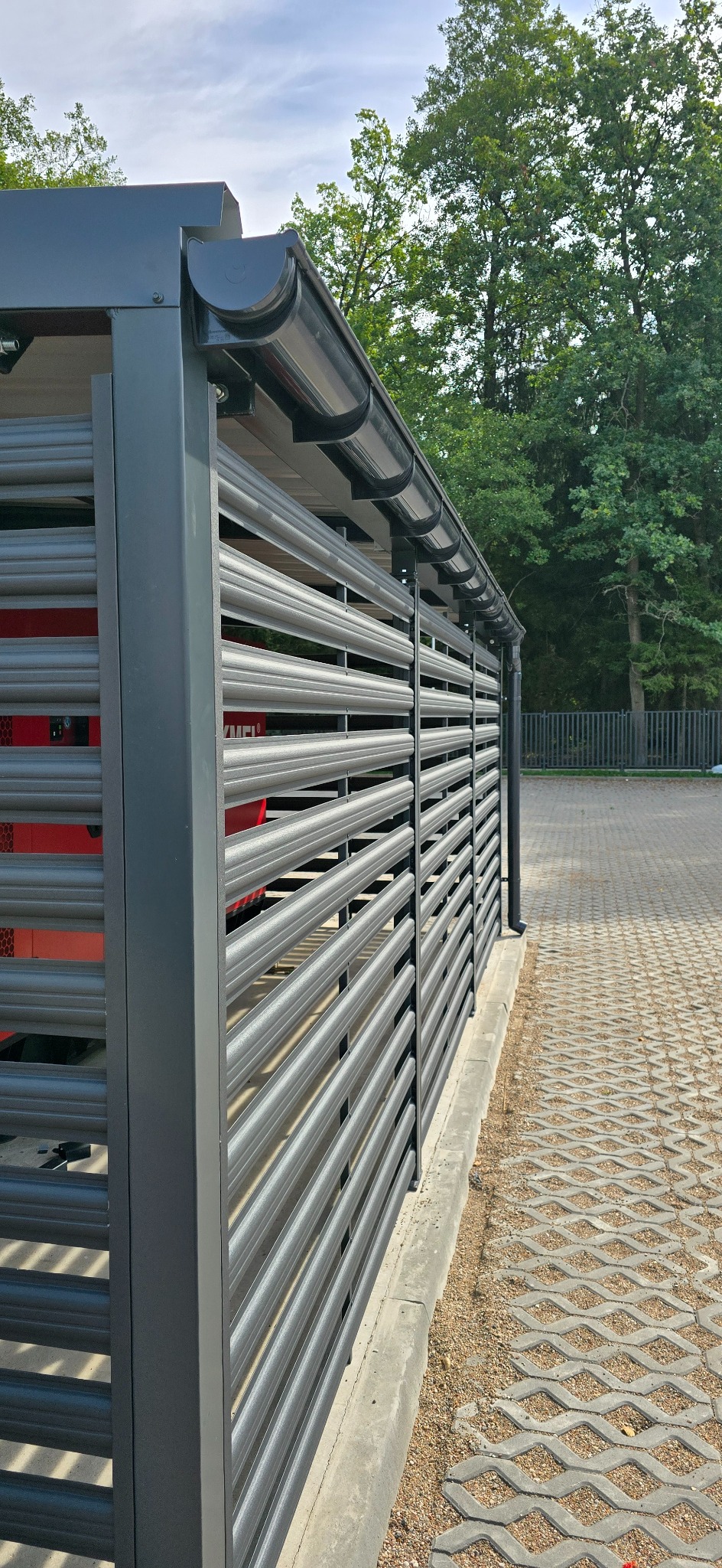 Nowoczesna, pozioma, metalowa konstrukcja z zadaszeniem i rynną, osłaniająca prawdopodobnie wiatę śmietnikową, z szarej blachy trapezowej, na tle kostki ażurowej i zieleni.