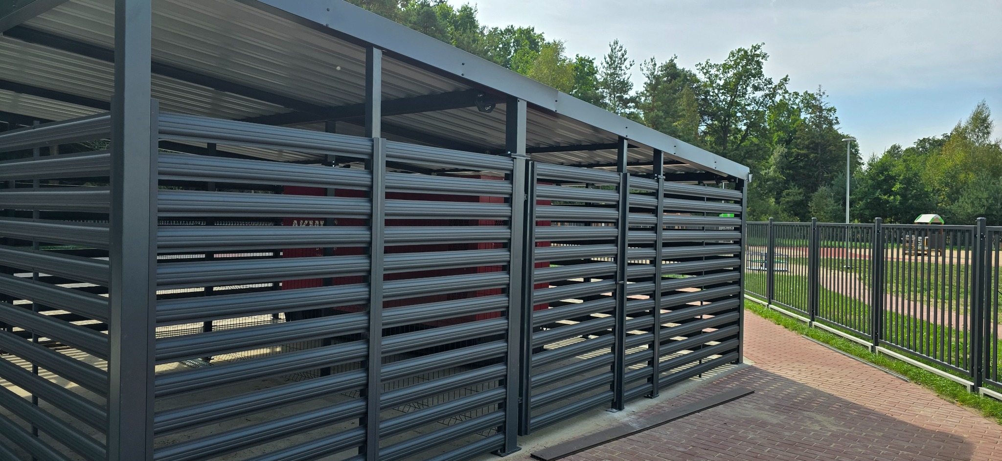 Metalowa konstrukcja zadaszona, z poziomymi panelami, prawdopodobnie osłona na śmietniki, widoczna z zewnątrz na tle zieleni i chodnika.