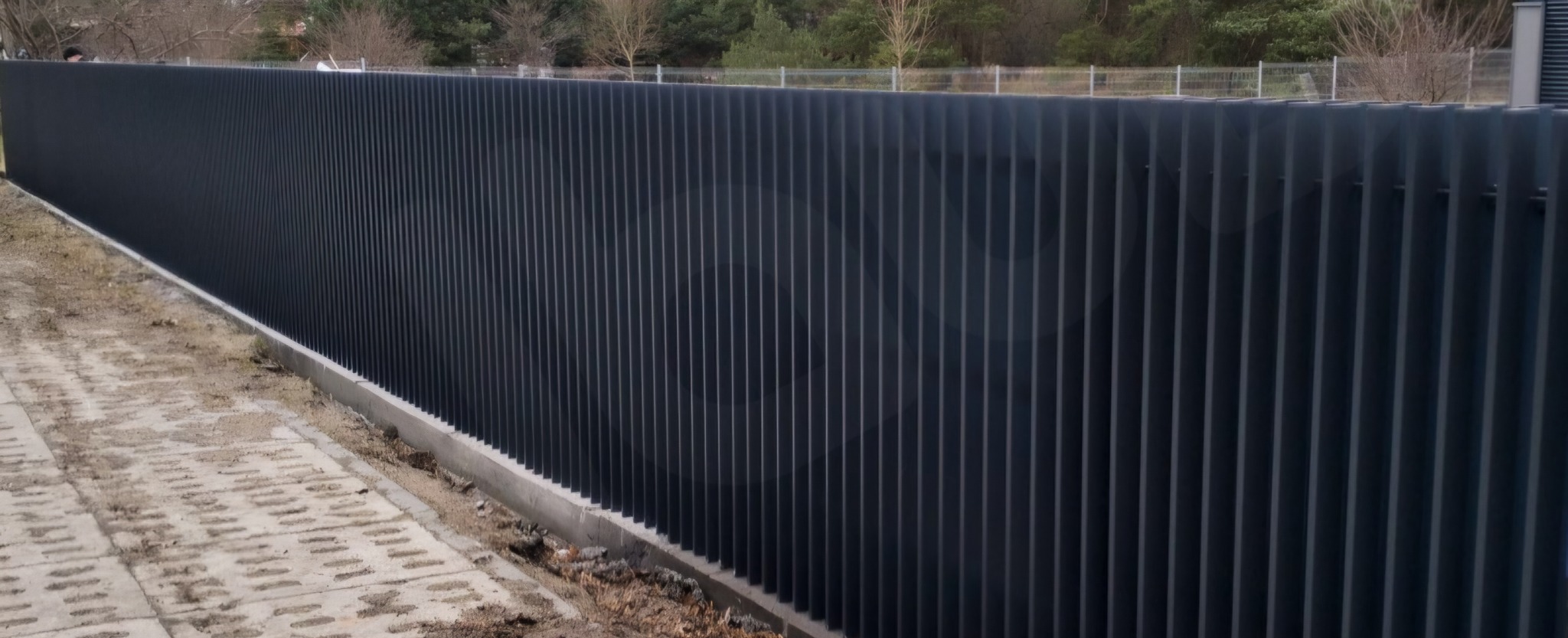 Długi, nowoczesny, ciemnoszary płot lamelowy z wąskich pionowych profili, oddzielający posesję od chodnika z betonowych płyt. W tle widoczne drzewa.