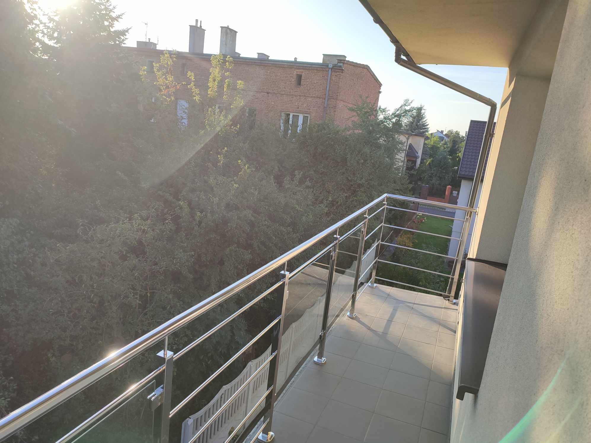 Balkon z nowoczesną balustradą ze stali nierdzewnej i szkła, widok na zieleń drzew i budynek z cegły w tle, płytki podłogowe w odcieniach szarości, poranne słońce.