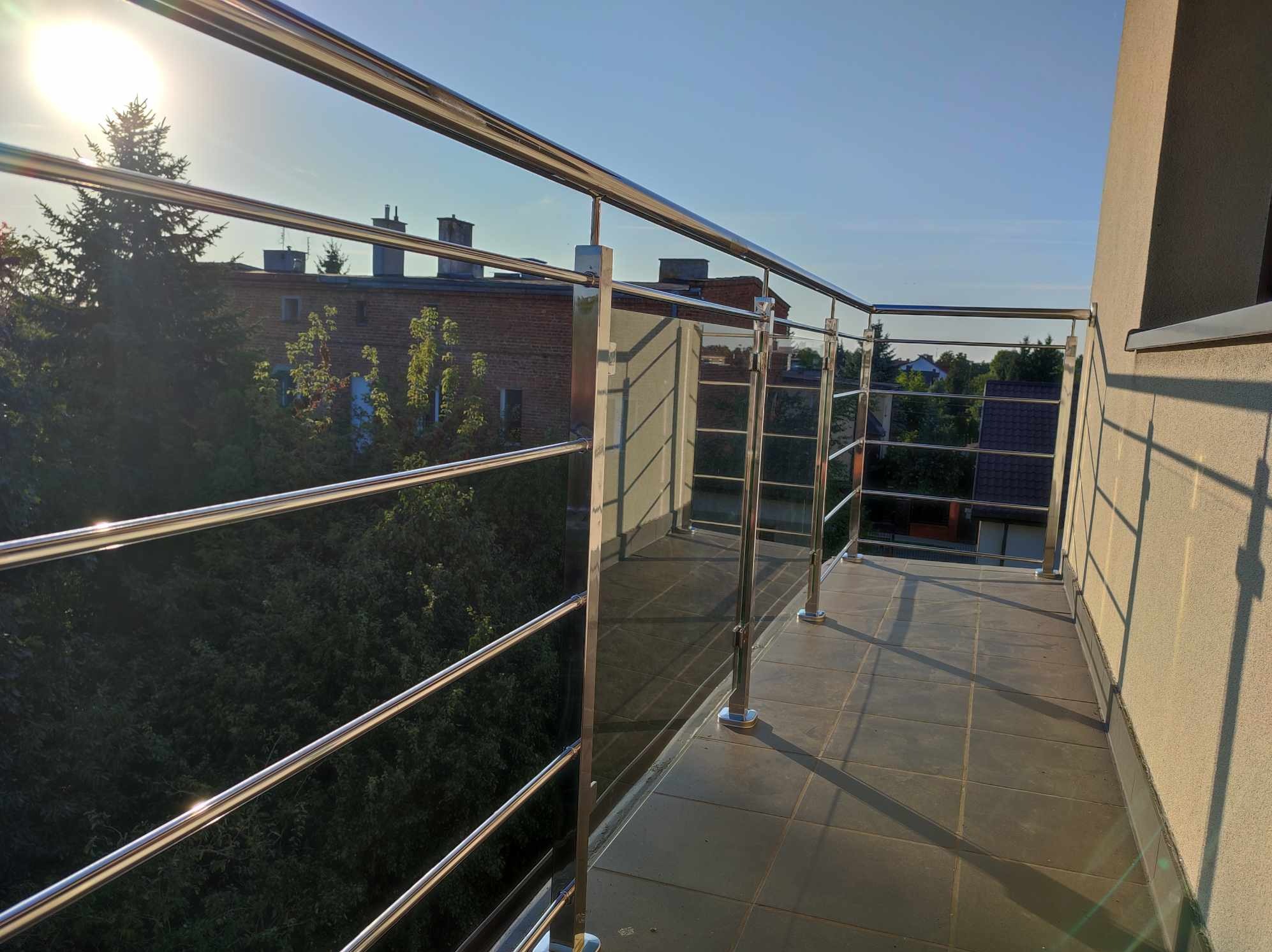 Błyszcząca, nowoczesna balustrada ze stali nierdzewnej na balkonie, z widokiem na zieleń i budynki w tle, uchwycona w promieniach popołudniowego słońca tworzących długie cienie na płytkach.