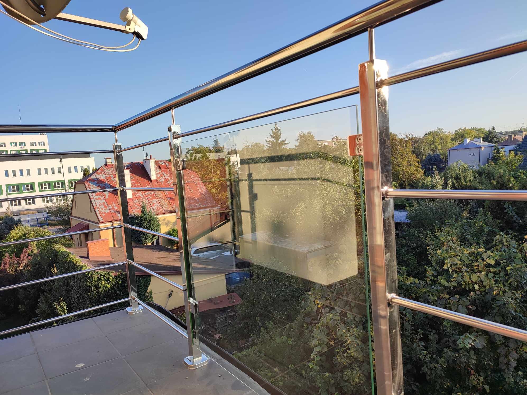 Balkon z nowoczesną balustradą ze stali nierdzewnej i szkła, widok na okoliczną zabudowę i zieleń, antena satelitarna w górnym rogu.