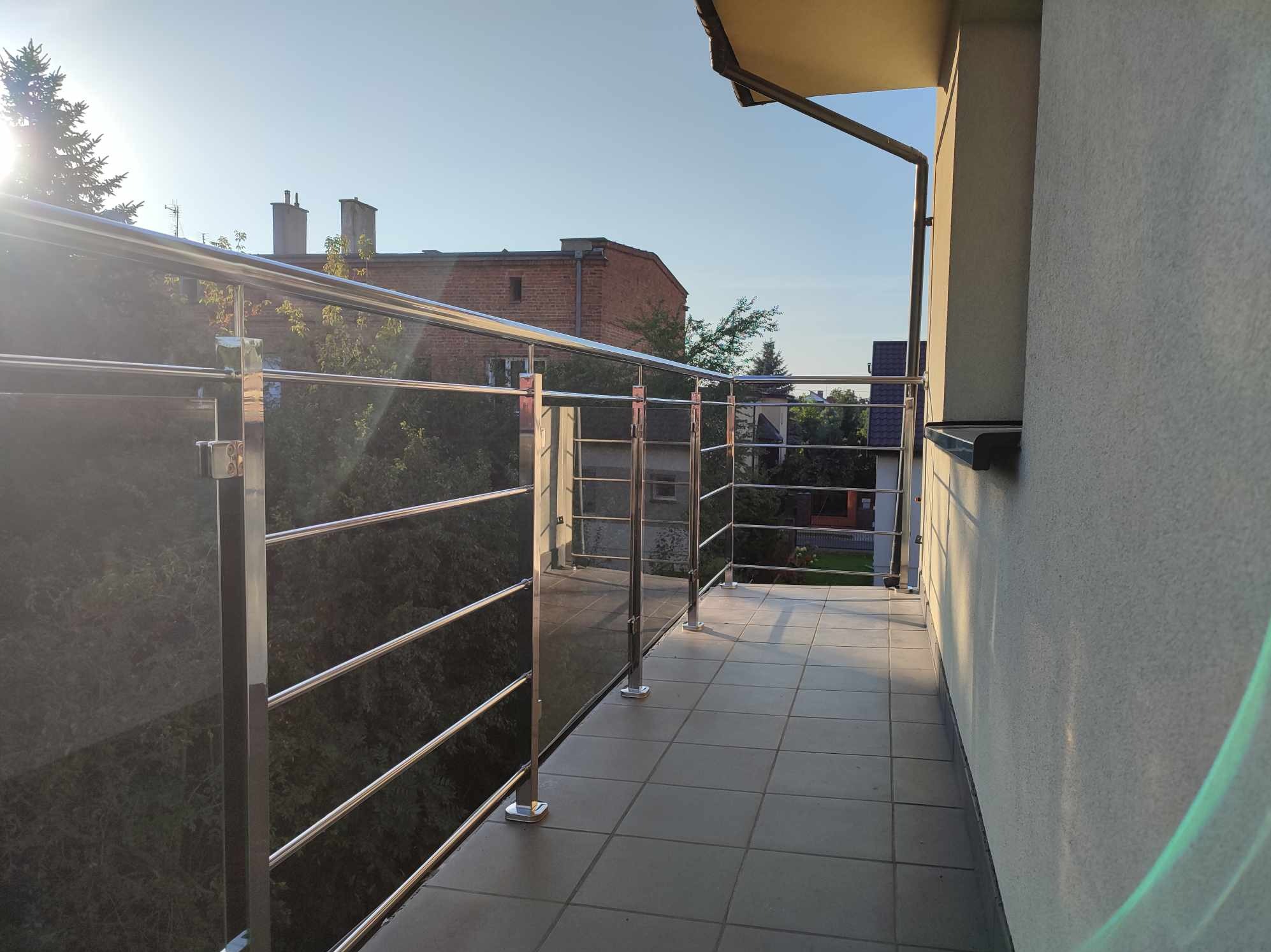 Balkon z balustradą ze stali nierdzewnej z czterema poziomymi prętami, widok na ceglany budynek i zieleń w tle, płytki na podłodze balkonu.