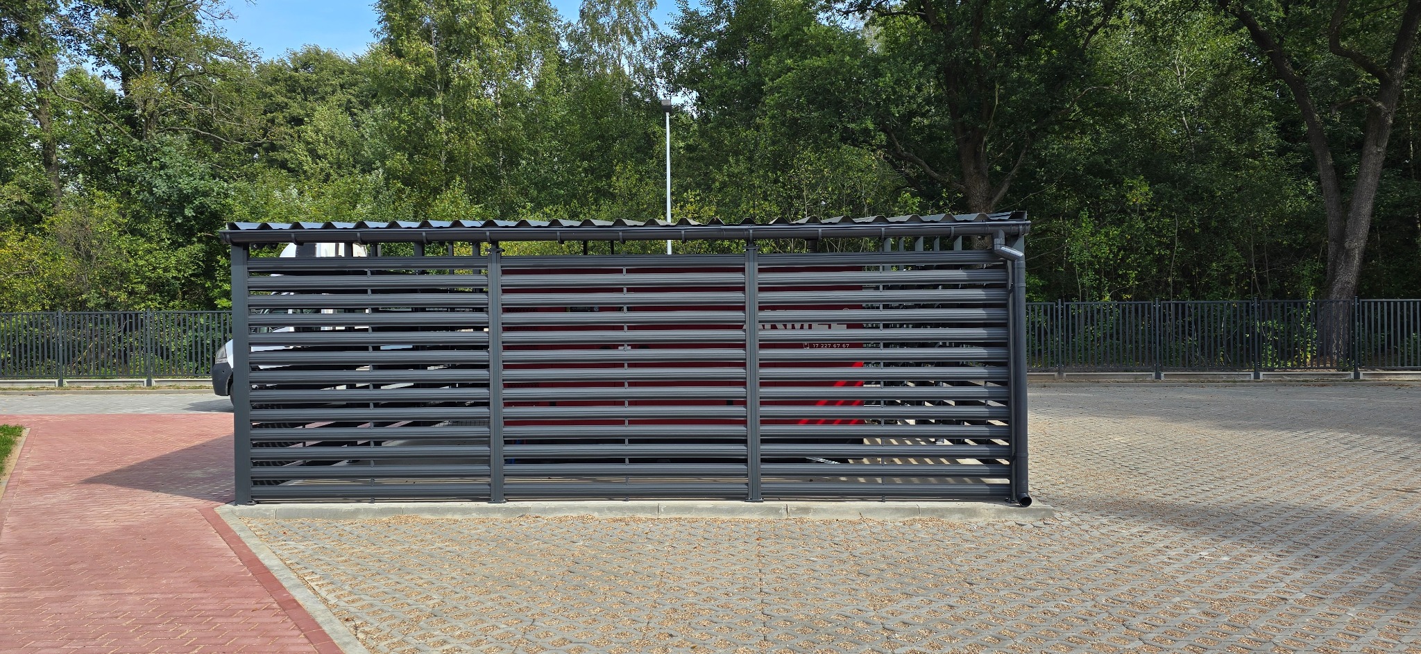 Szara, metalowa konstrukcja z poziomymi szczeblami i dachem, prawdopodobnie wiata śmietnikowa, na tle drzew i parkingu z ażurowych płyt.