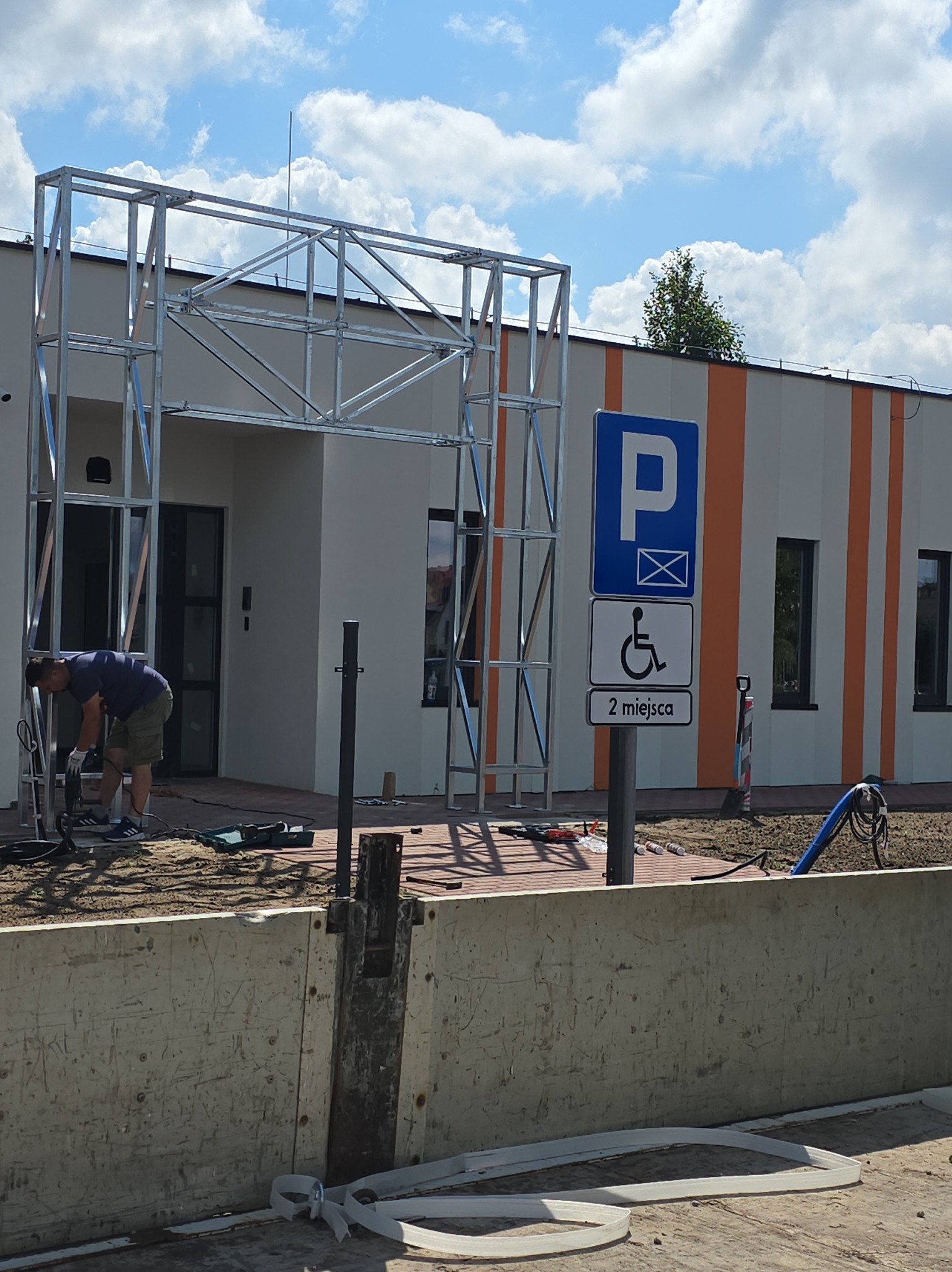 Pracownik w Łukowie montuje metalową konstrukcję przy budynku z oznaczeniem parkingu dla niepełnosprawnych, widoczne narzędzia pracy.