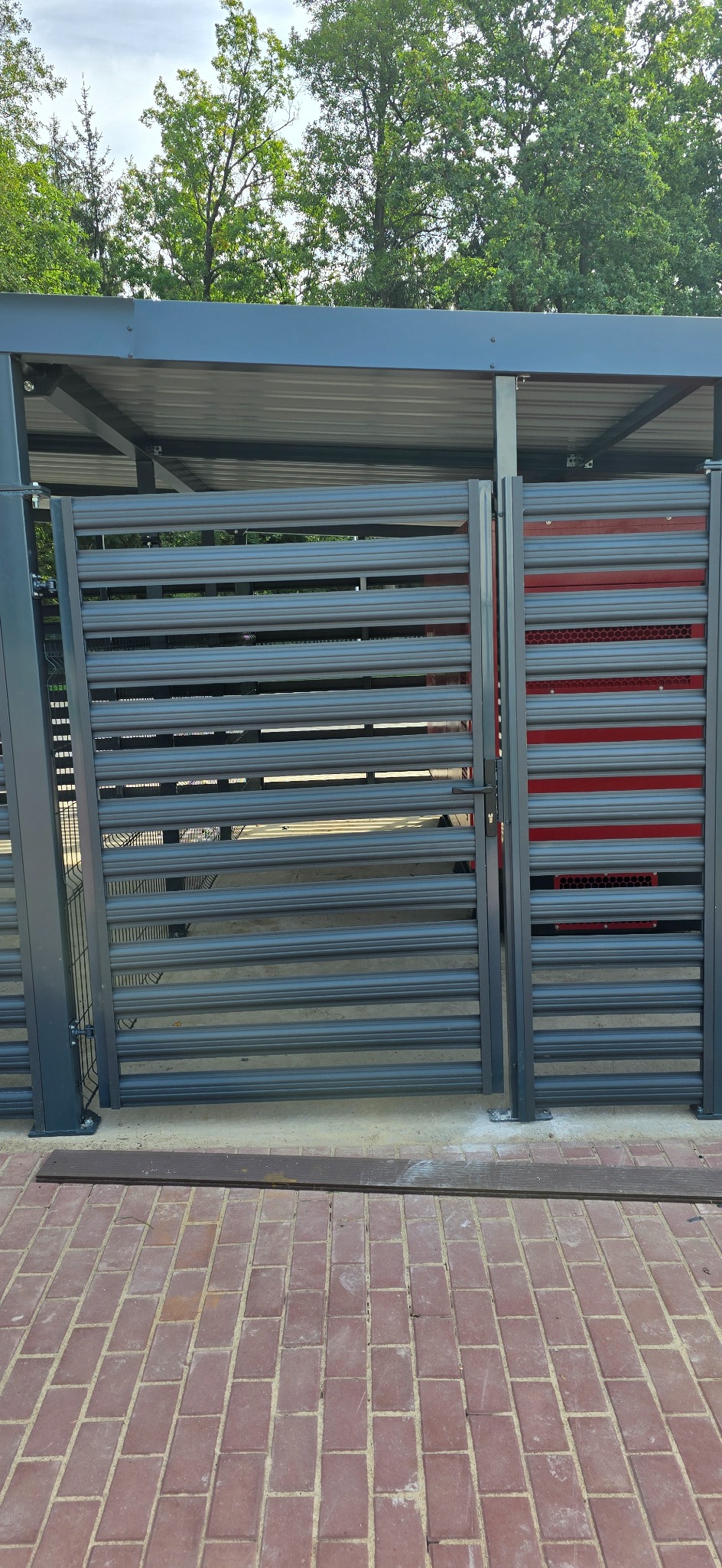 Nowoczesna, pozioma, szara brama lamelowa z widocznym fragmentem czerwonego kontenera na śmieci w tle, umieszczona pod zadaszeniem, na tle ceglanej kostki brukowej.