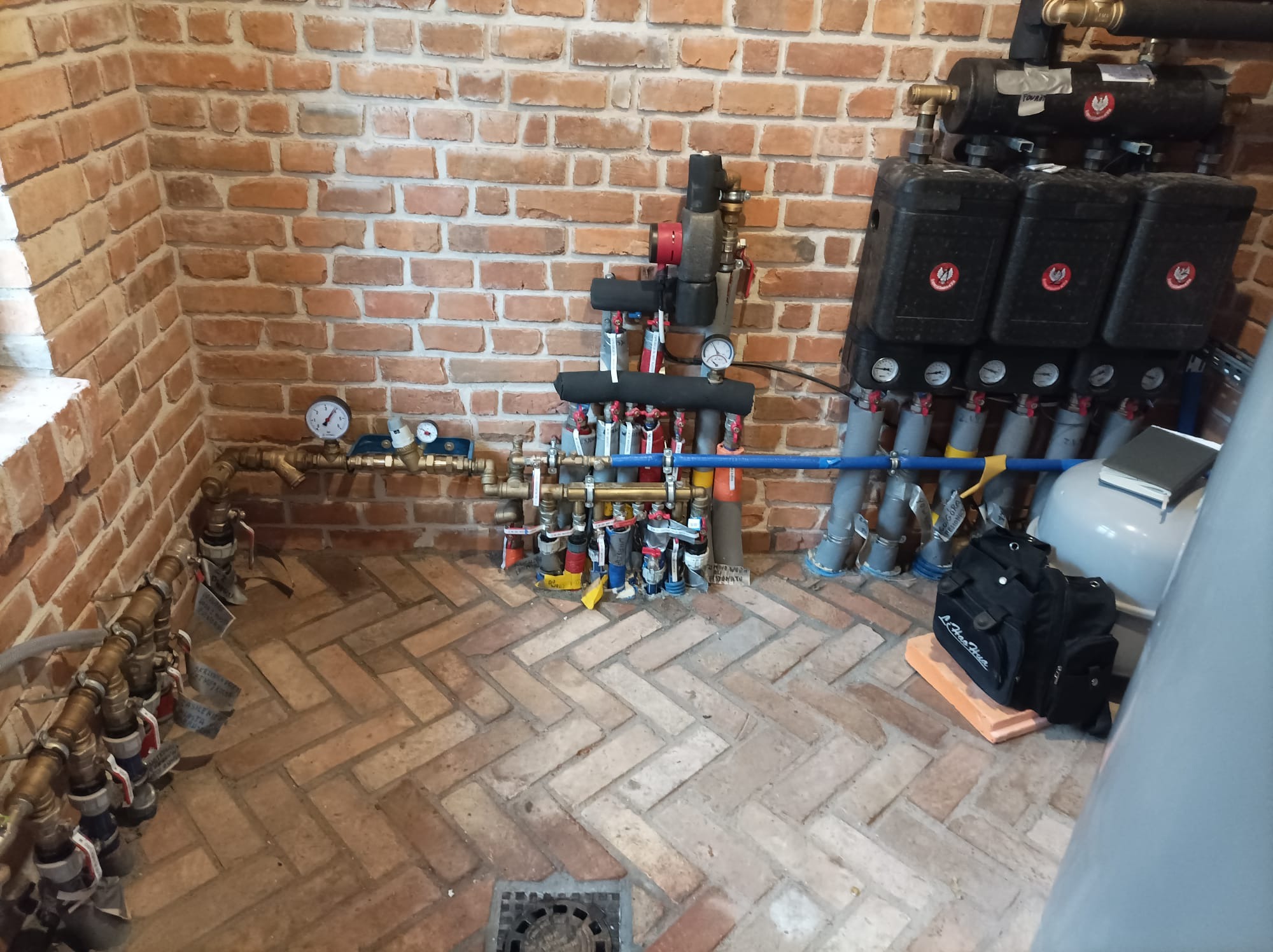 Rozdzielacz ogrzewania podłogowego z pompą obiegową i grupą pompową, zamontowany w pomieszczeniu z ceglanymi ścianami i podłogą ułożoną w jodełkę, widoczny również czarny worek narzędziowy.