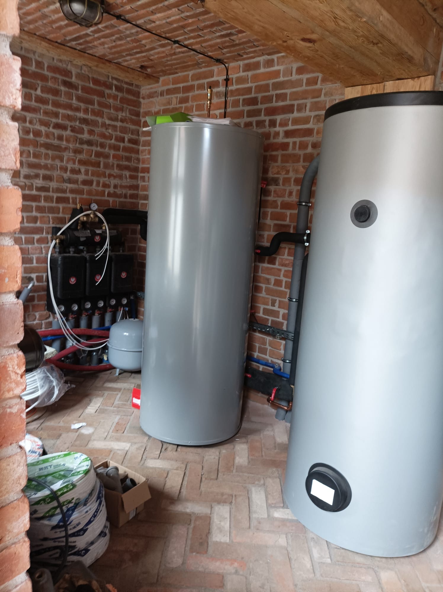 Dwa szare zbiorniki akumulacyjne ciepła w kotłowni z ceglanymi ścianami i drewnianym stropem, widoczna instalacja hydrauliczna z czarnymi i czerwonymi rurami, na podłodze resztki materiałów...
