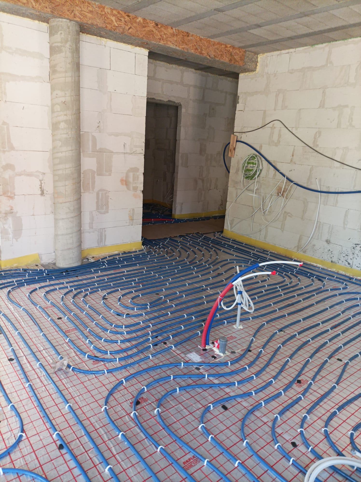 Niebieskie rurki ogrzewania podłogowego ułożone na folii z czerwoną siatką w surowym pomieszczeniu z betonowych pustaków, widoczne białe uchwyty mocujące i wystające rury przyłączeniowe.
