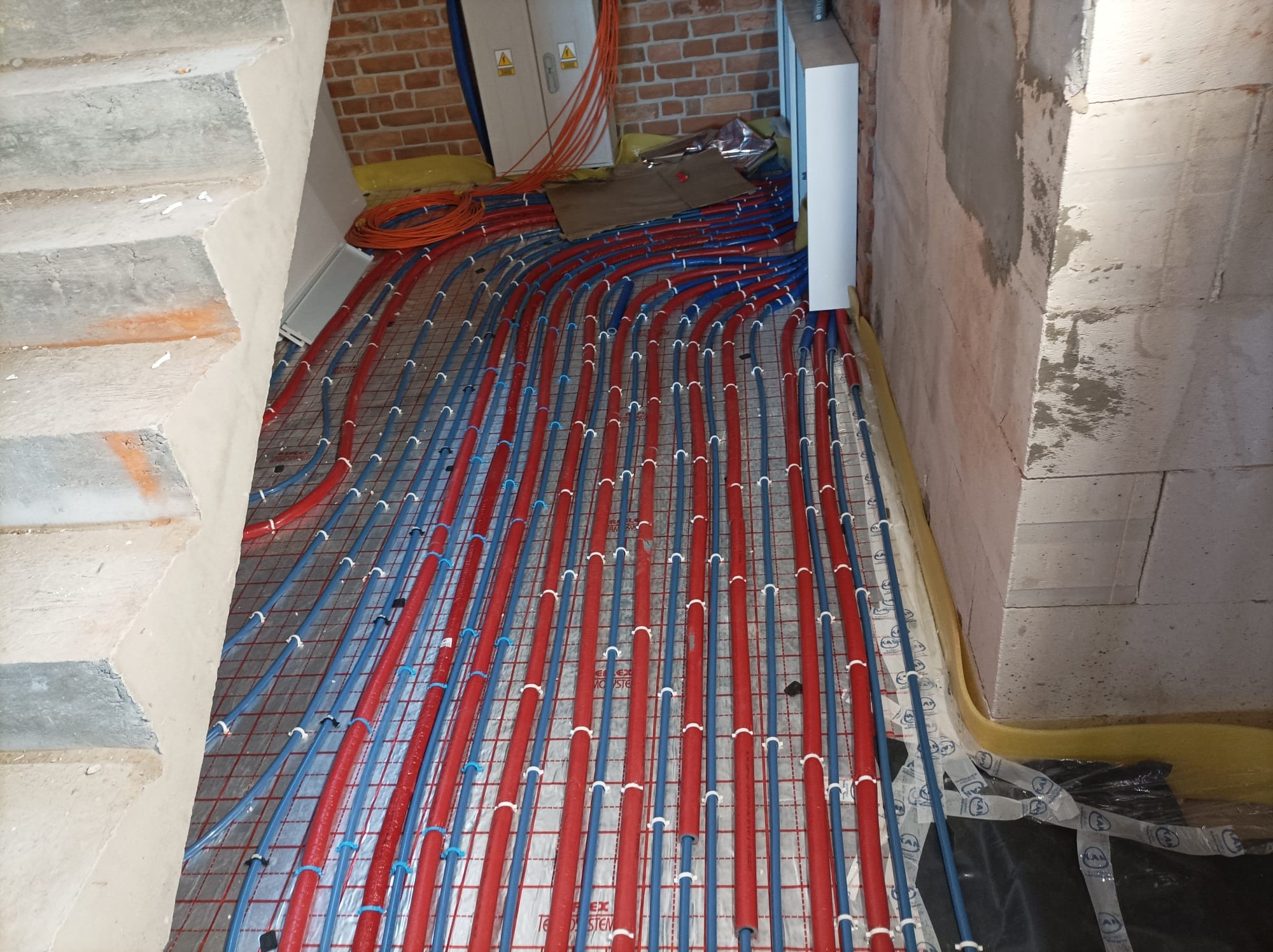 Rozłożona instalacja ogrzewania podłogowego z czerwonymi i niebieskimi rurami na siatce, widoczne elementy elektryczne na ścianie z cegły, surowy stan wykończenia.