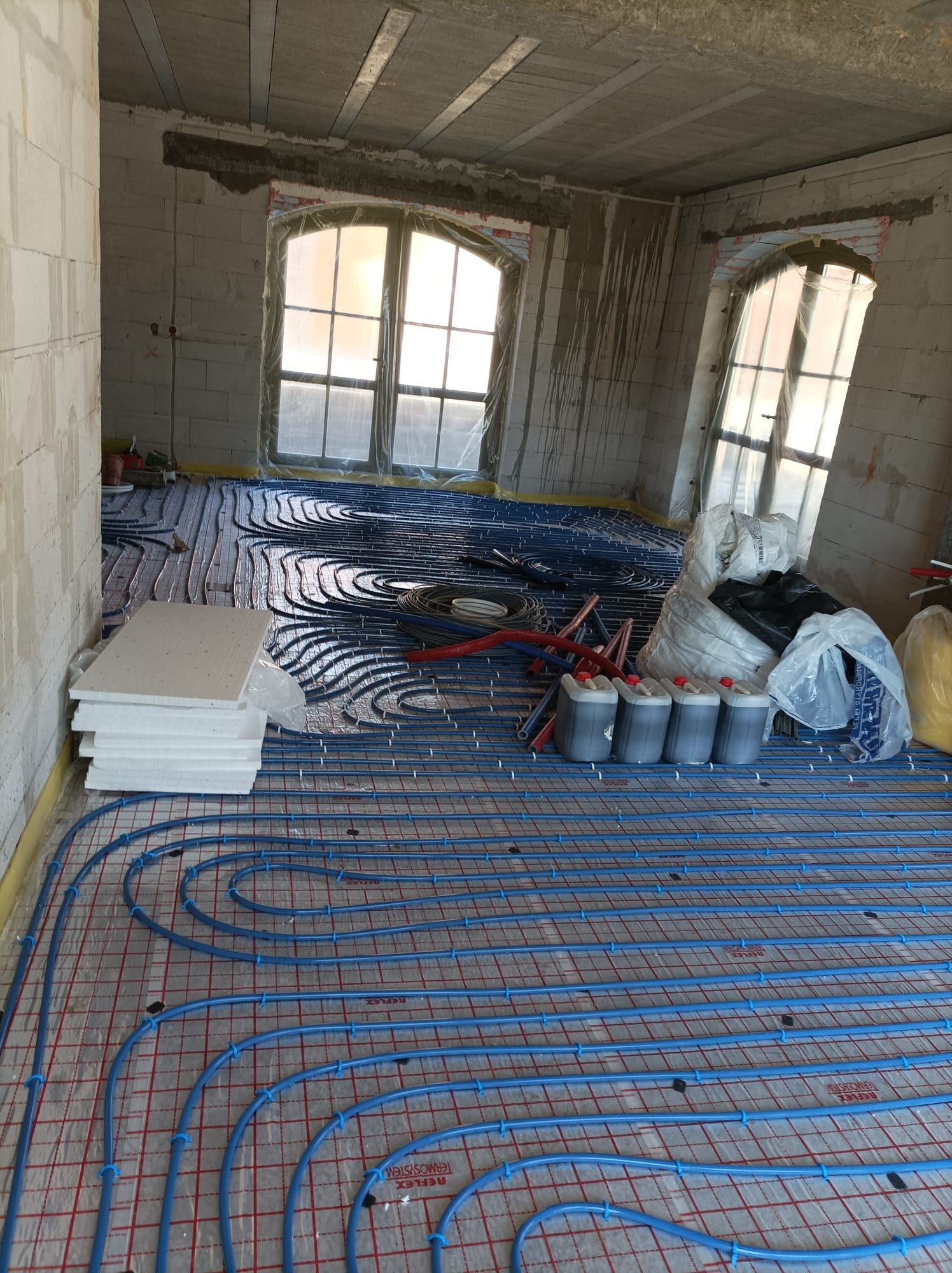 Niedokończona instalacja ogrzewania podłogowego w surowym stanie, widoczne rury w kolorze niebieskim ułożone na czerwonej siatce, styropian izolacyjny i pojemniki z płynem do ogrzewania, okna...