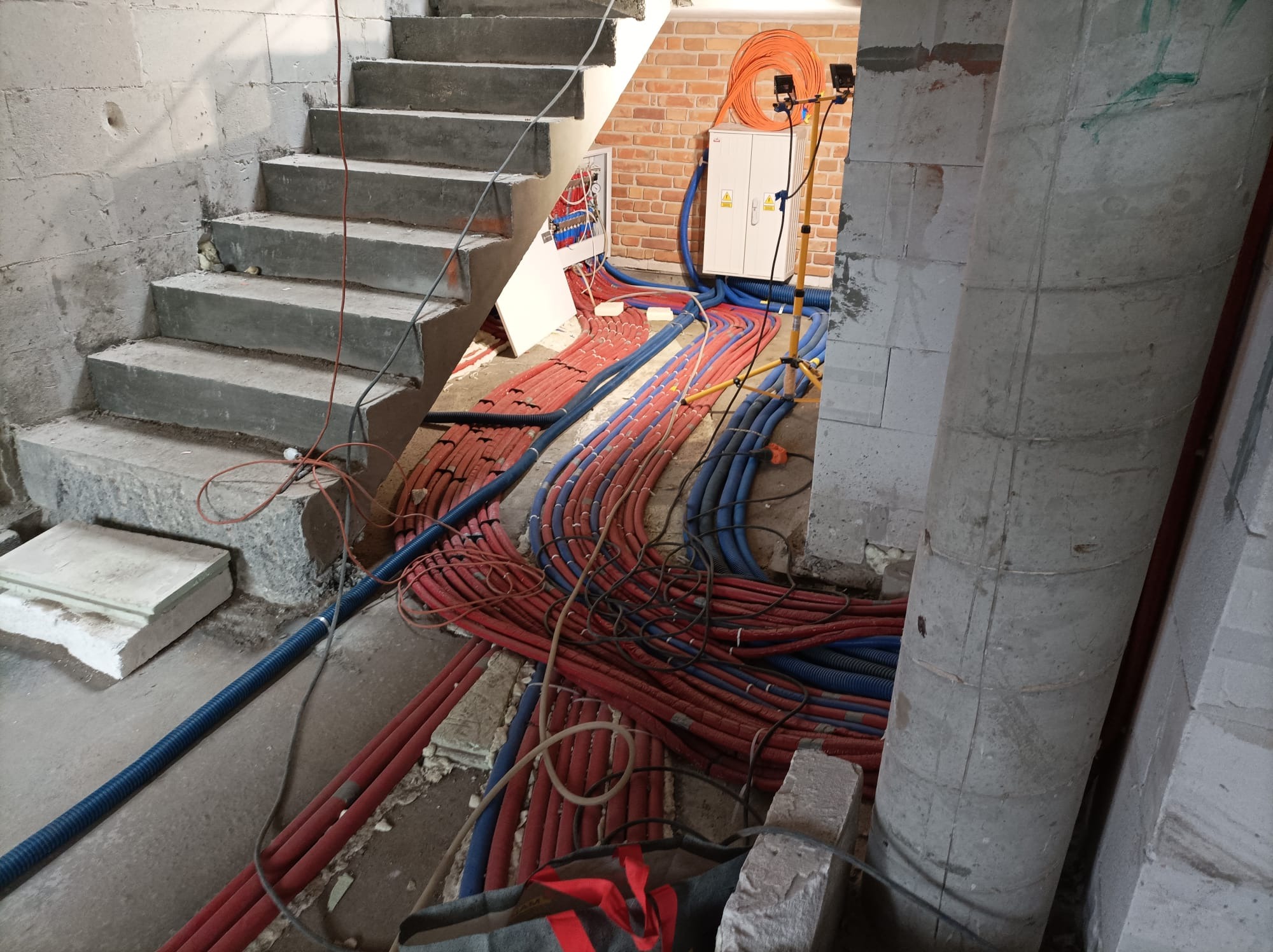 Instalacja elektryczna i hydrauliczna w surowym stanie pod schodami: widoczne liczne czerwone i niebieskie rury, skrzynka elektryczna na ceglanej ścianie, betonowe schody i słup.