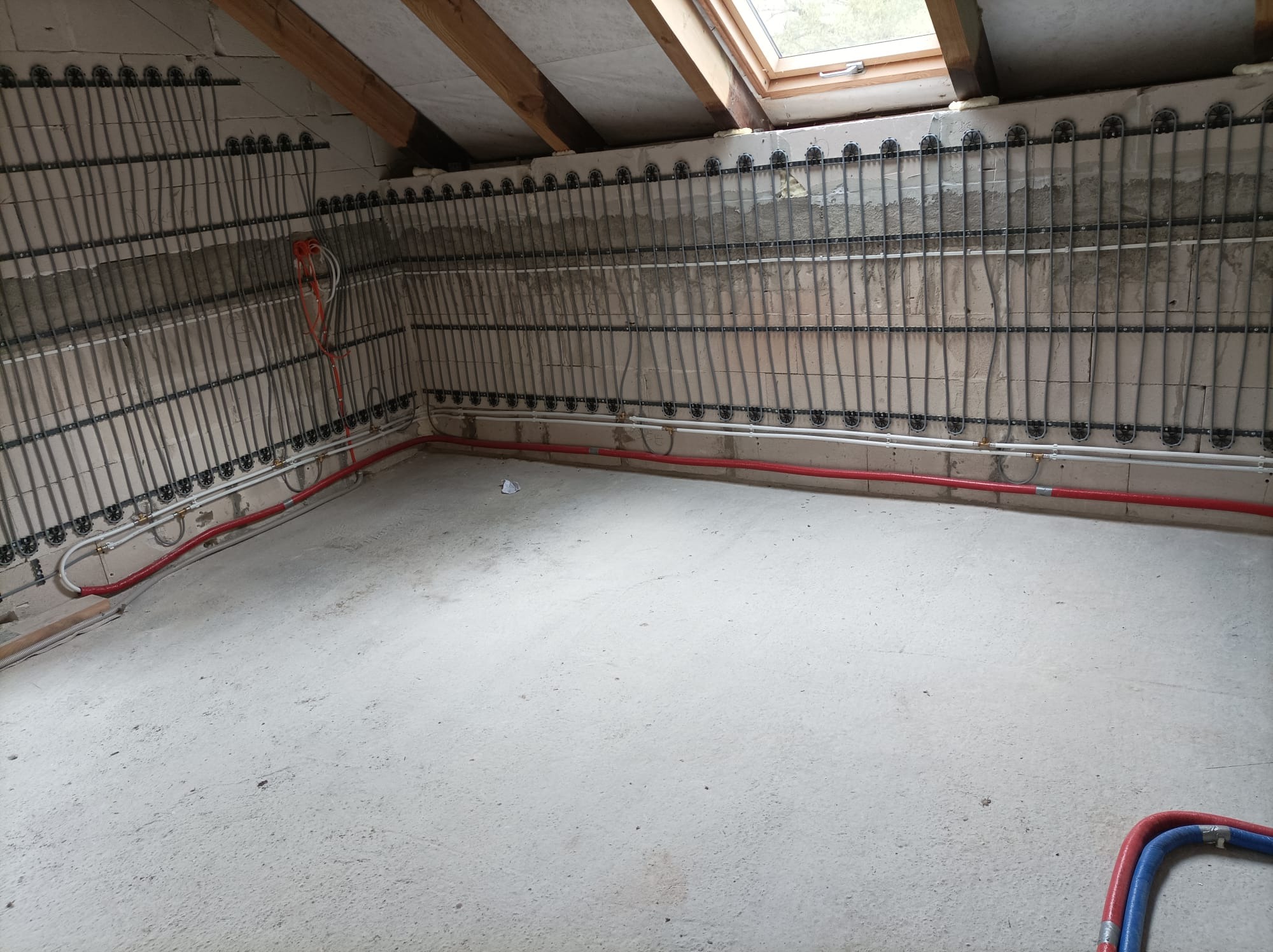 Montaż rur grzewczych na ścianie w pomieszczeniu ze skosem, widoczne elementy mocujące, czerwone i białe rury biegnące wzdłuż ściany, surowy beton na podłodze i ścianach, okno dachowe.