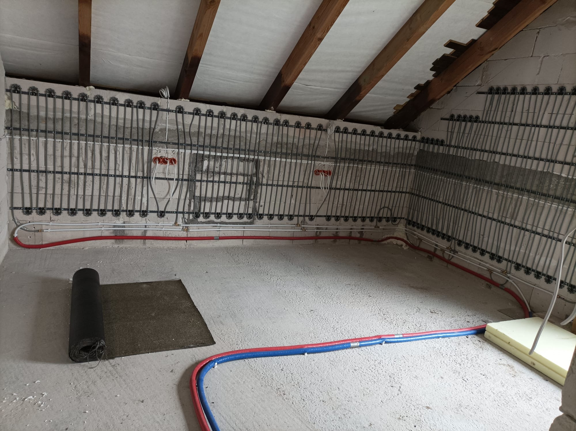 Ściany poddasza z zamontowaną instalacją ogrzewania podłogowego ściennego, rurki rozprowadzone w pionowych liniach, widoczne również rury doprowadzające wodę, na podłodze leży zwinięta mata...