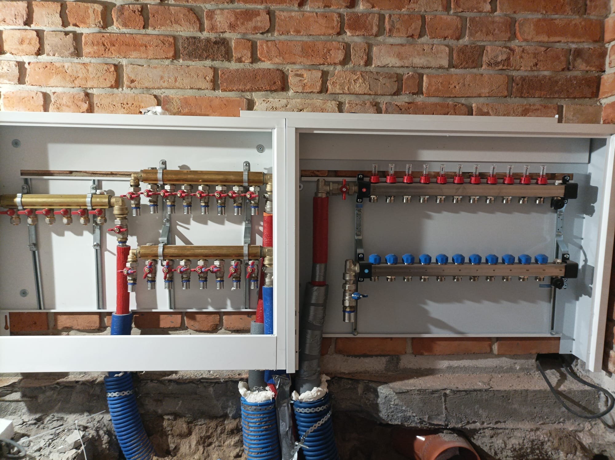 Rozdzielacz instalacji grzewczej podłogowej w szafce, z widocznymi rurami doprowadzającymi i ceglaną ścianą w tle. Część instalacji zabezpieczona pianką montażową.