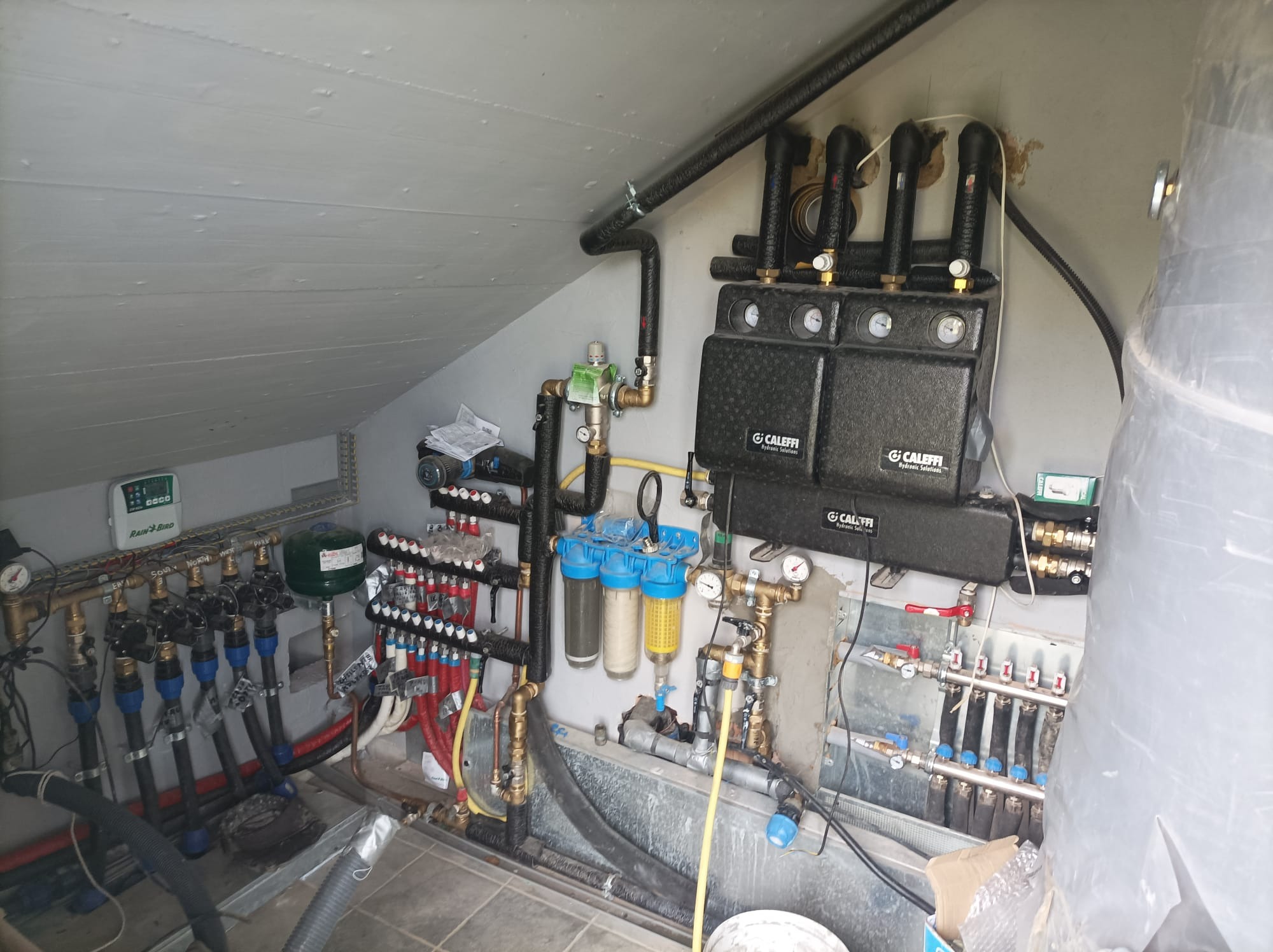 Rozdzielacz ogrzewania podłogowego z filtrami i elementami sterującymi, zainstalowany w Szczecinku, widoczny w skośnym pomieszczeniu z drewnianą ścianą.