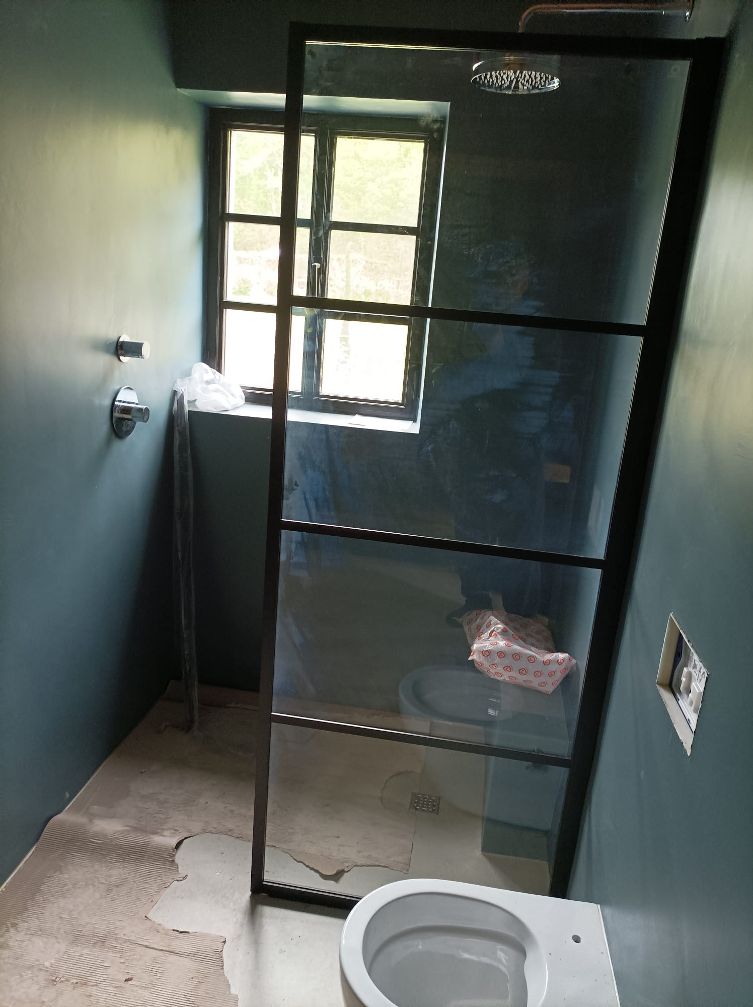 Wnętrze łazienki w trakcie remontu z kabiną prysznicową w stylu loft, widoczne okno z czarną ramą, biała toaleta i fragmenty uszkodzonej podłogi.