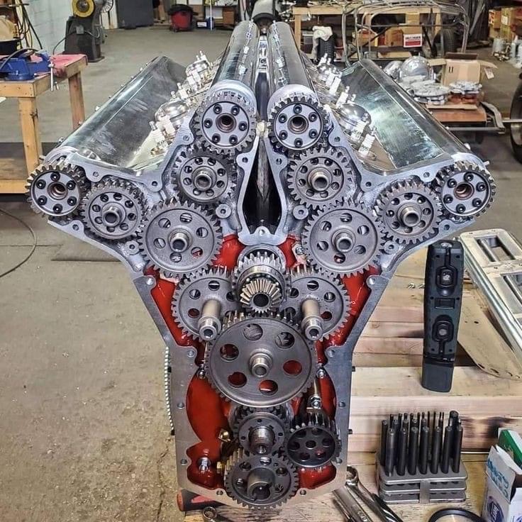Widok z przodu silnika V-kształtnego z widocznymi licznymi zębatkami i czerwonym wnętrzem bloku, narzędzia warsztatowe leżą obok.